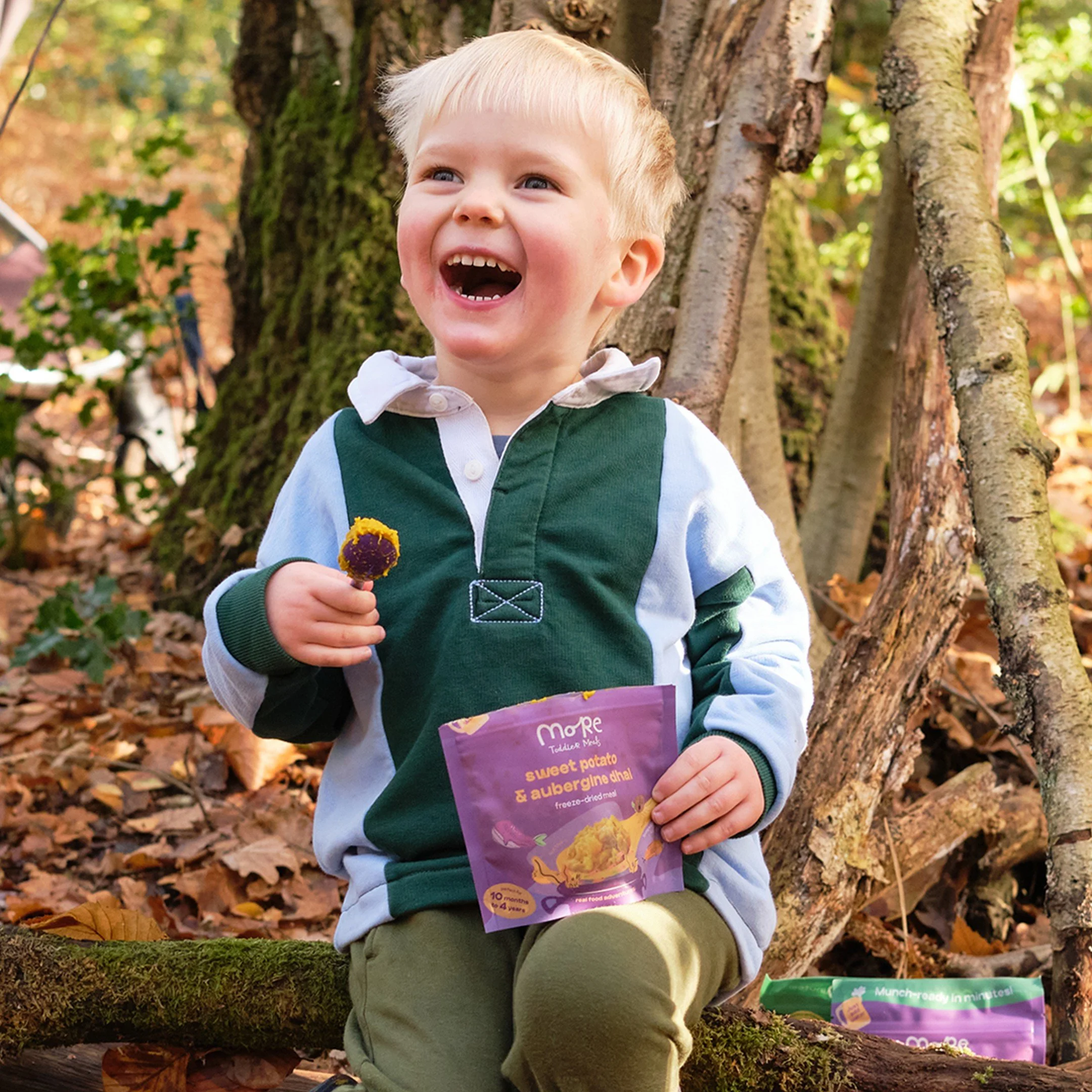 Smiling young child holding a purple packet of More Toddler Foods and sitting on a fallen tree in a woodland setting.