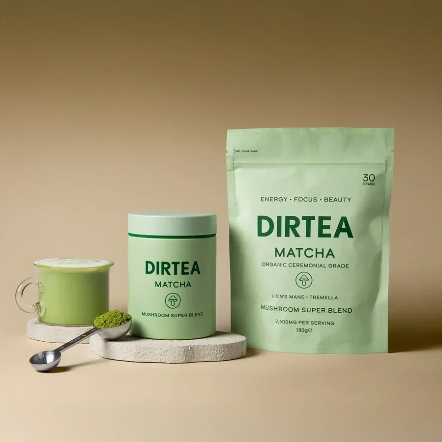 A glass of matcha tea next to a green tub and a pouch of DIRTEA Matcha Mushroom Super Blend