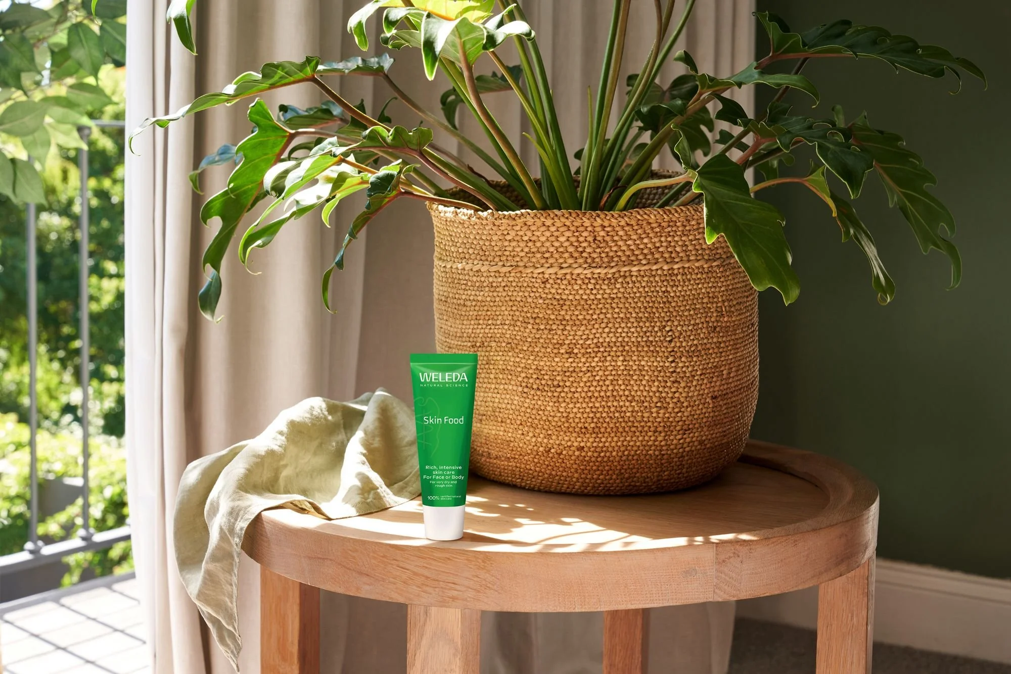 Weleda Skin Food tube on a wooden table with a green plant in a weaved basket in the background.