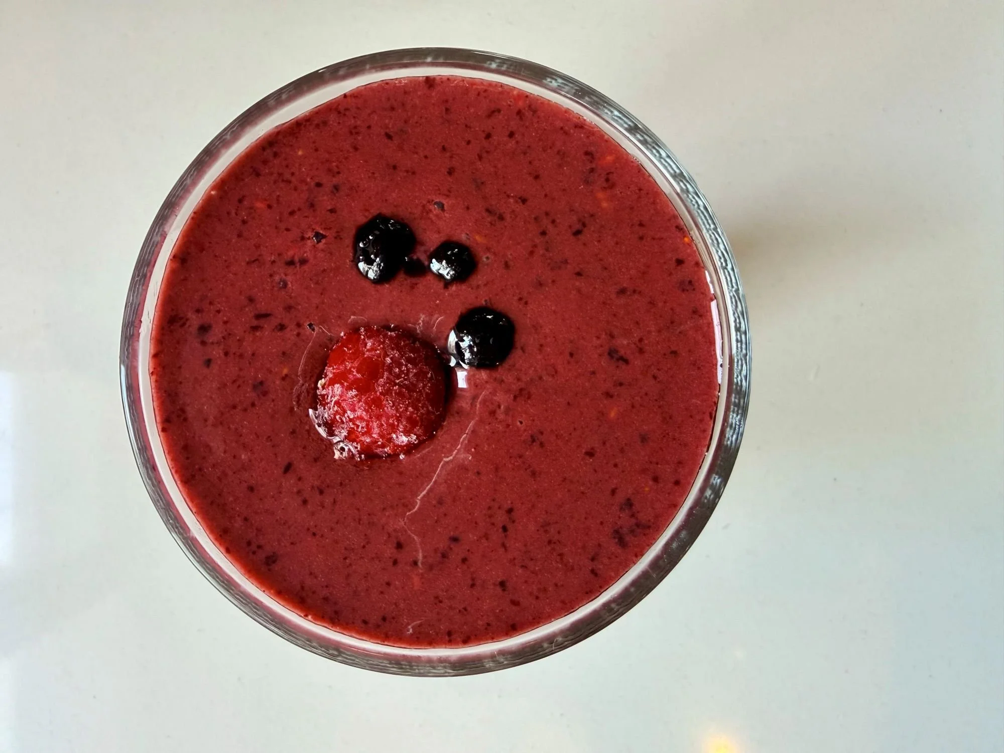 A top-down view of a deep red berry smoothie in a clear glass, with a raspberry and three blueberries arranged on the surface against a plain light background.