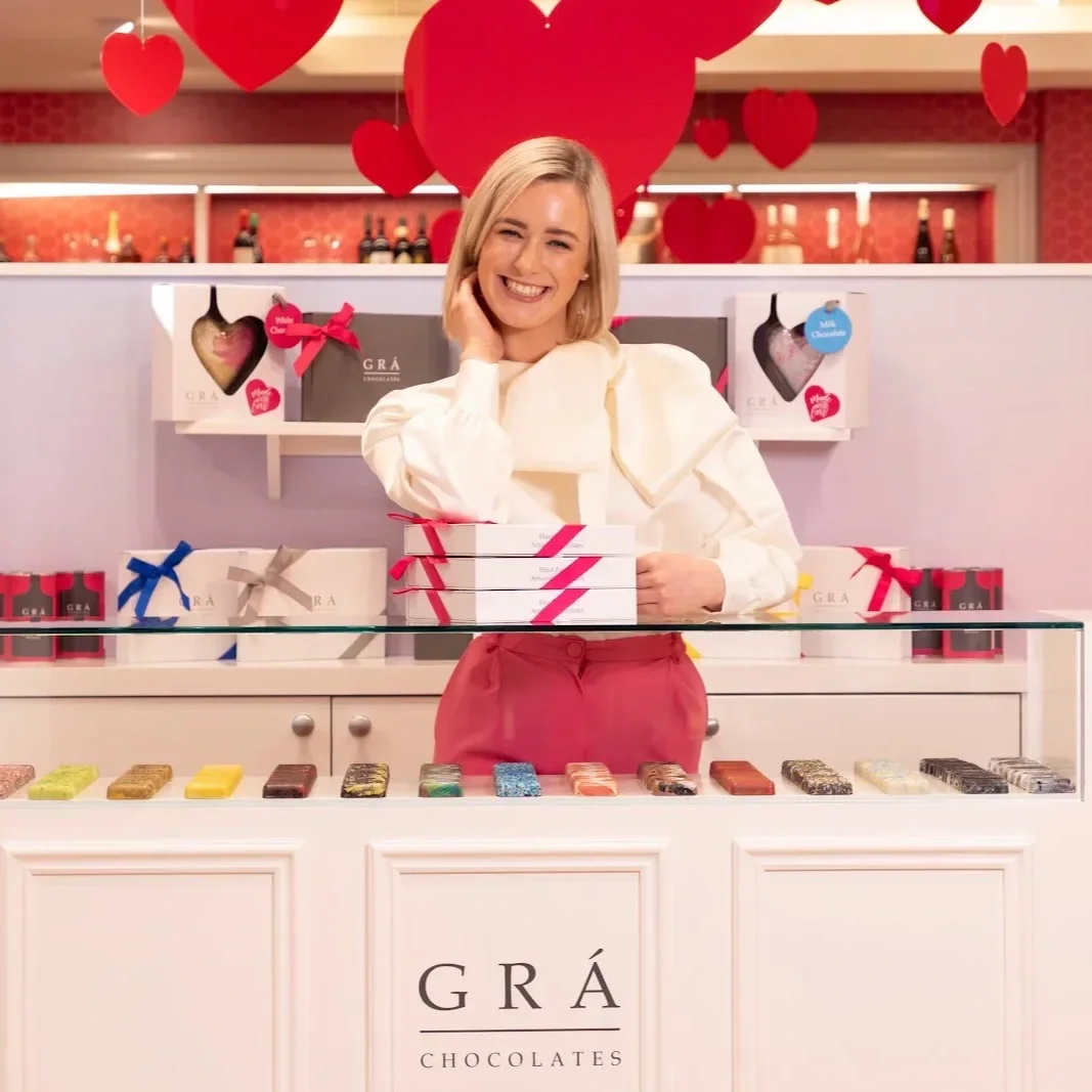 Woman standing behind the counter in a GRÁ Chocolates shop, with boxed chocolates on display and red heart decorations hanging overhead.