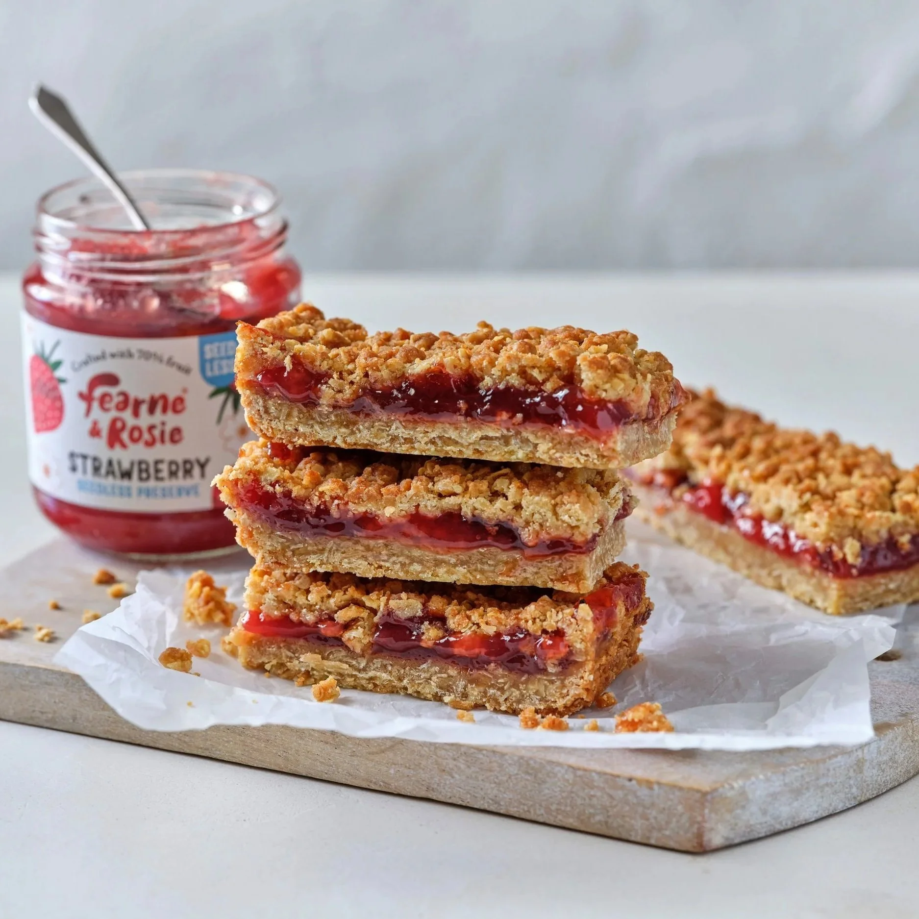 Stack of jam crumble bars on a wooden board next to an open jar of Fearne and Rosie strawberry preserve and spoon.