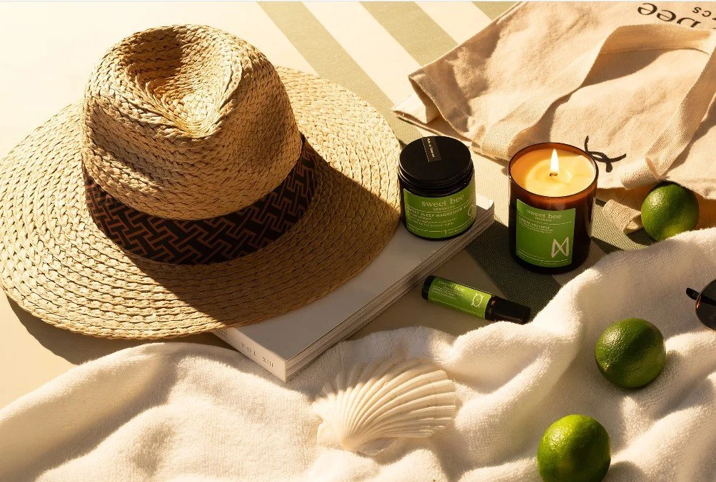 Sweet Bee Organics products on a white beach towel along with a straw hat, book, limes and lit candle