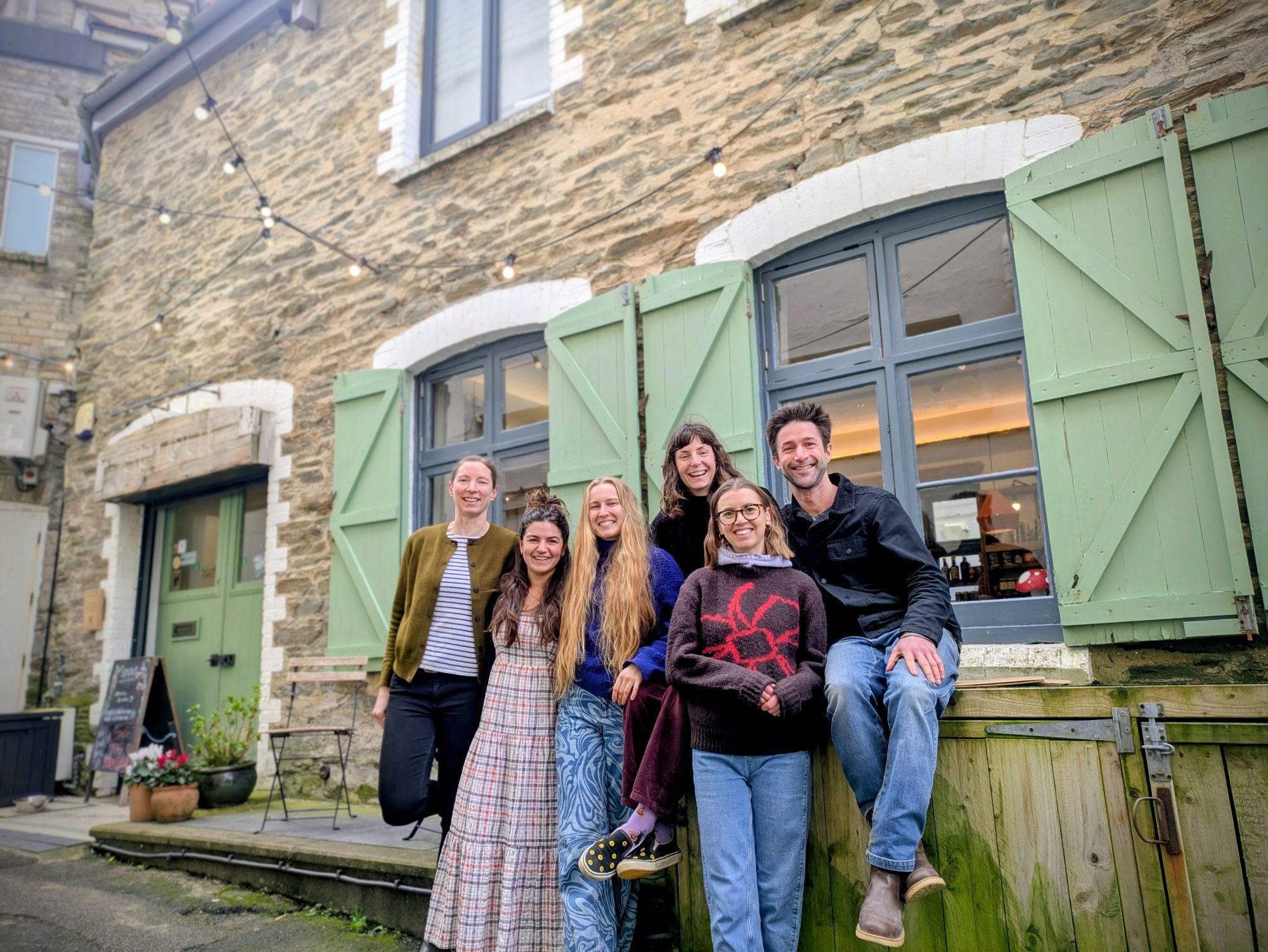 Spout Health Foods owners Sebastian and Claire Venn pose with four employees outside a rustic stone building with sage-green shutters.
