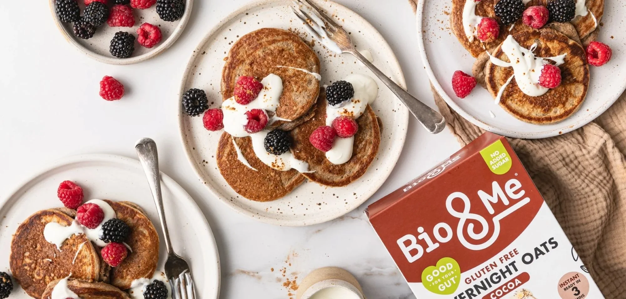 Plates of small pancakes topped with yogurt and berries, with a box of Bio&Me gluten-free overnight oats nearby.