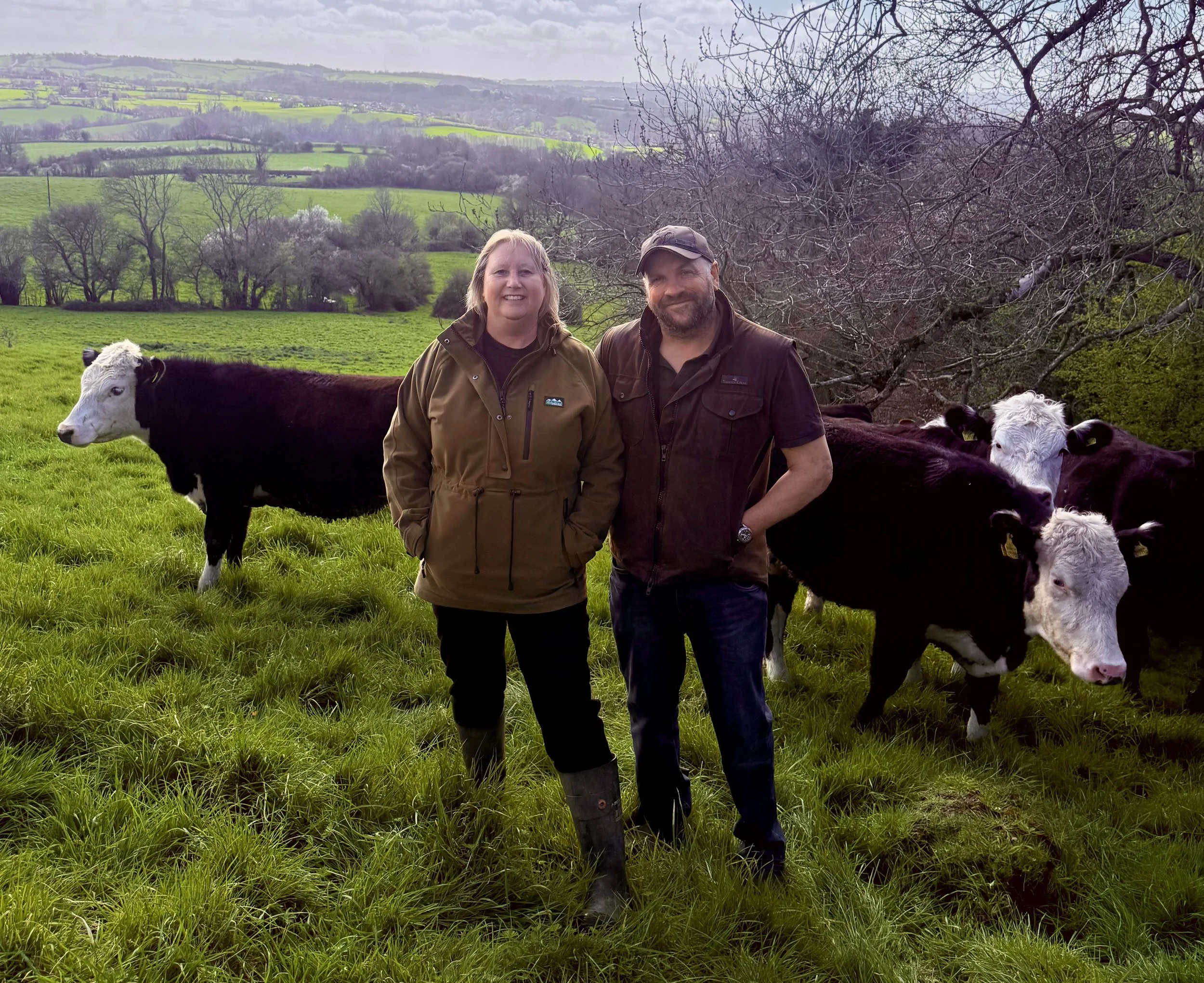 Teresa and Charlie Allward standing in a field with three black and white cows behind them.