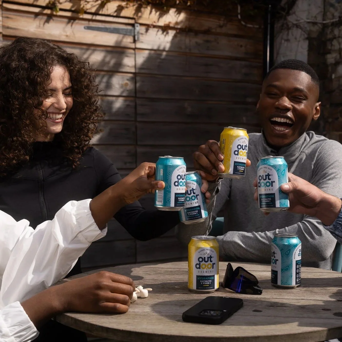 Group of four people sitting around an outdoor table, smiling and clinking cans of drinks together in a casual garden setting, with wooden fencing and a brick wall in the background.