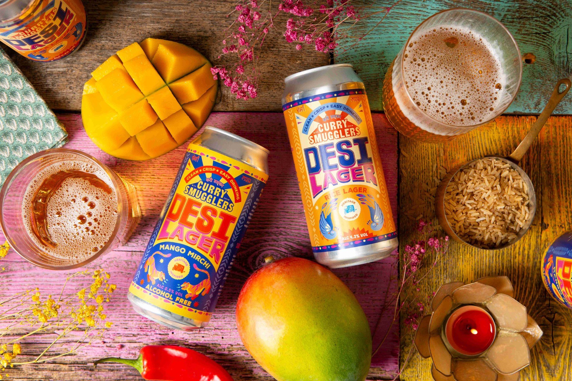 View from above of 2 Desi Lager cans on a table with mangos, glasses of beer, chillis and rice grains