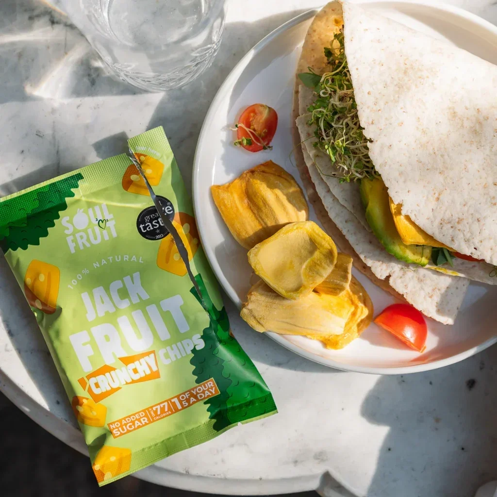 Pack of Soul Fruit Jackfruit Crunchy Chips displayed next to a wrap, dried jackfruit and cherry tomatoes on a plate