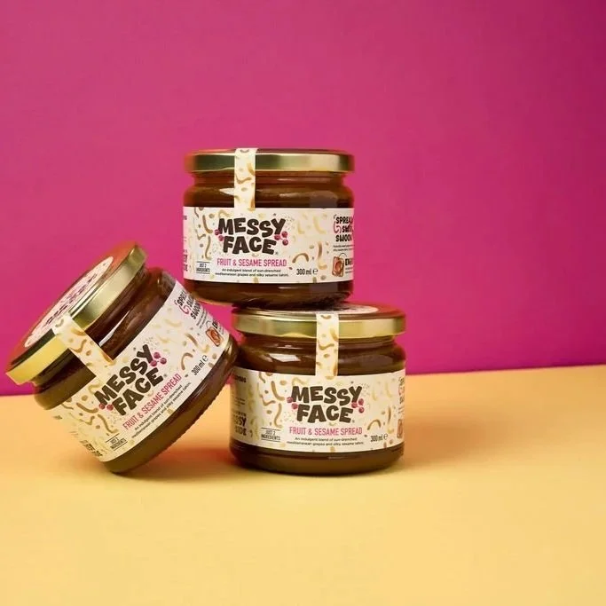 Three jars of Messy Face fruit and sesame spread piled up against a bright pink background.