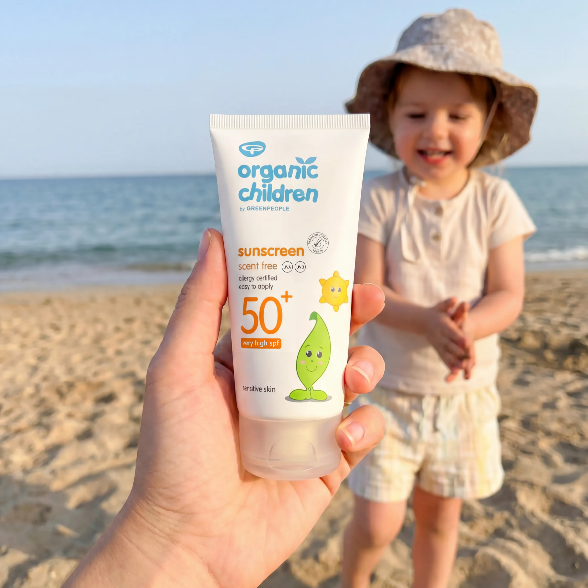 Hand holding a Green People Organic Children SPF50+ sunscreen tube on a beach, with a young child in a sun hat blurred in the background.