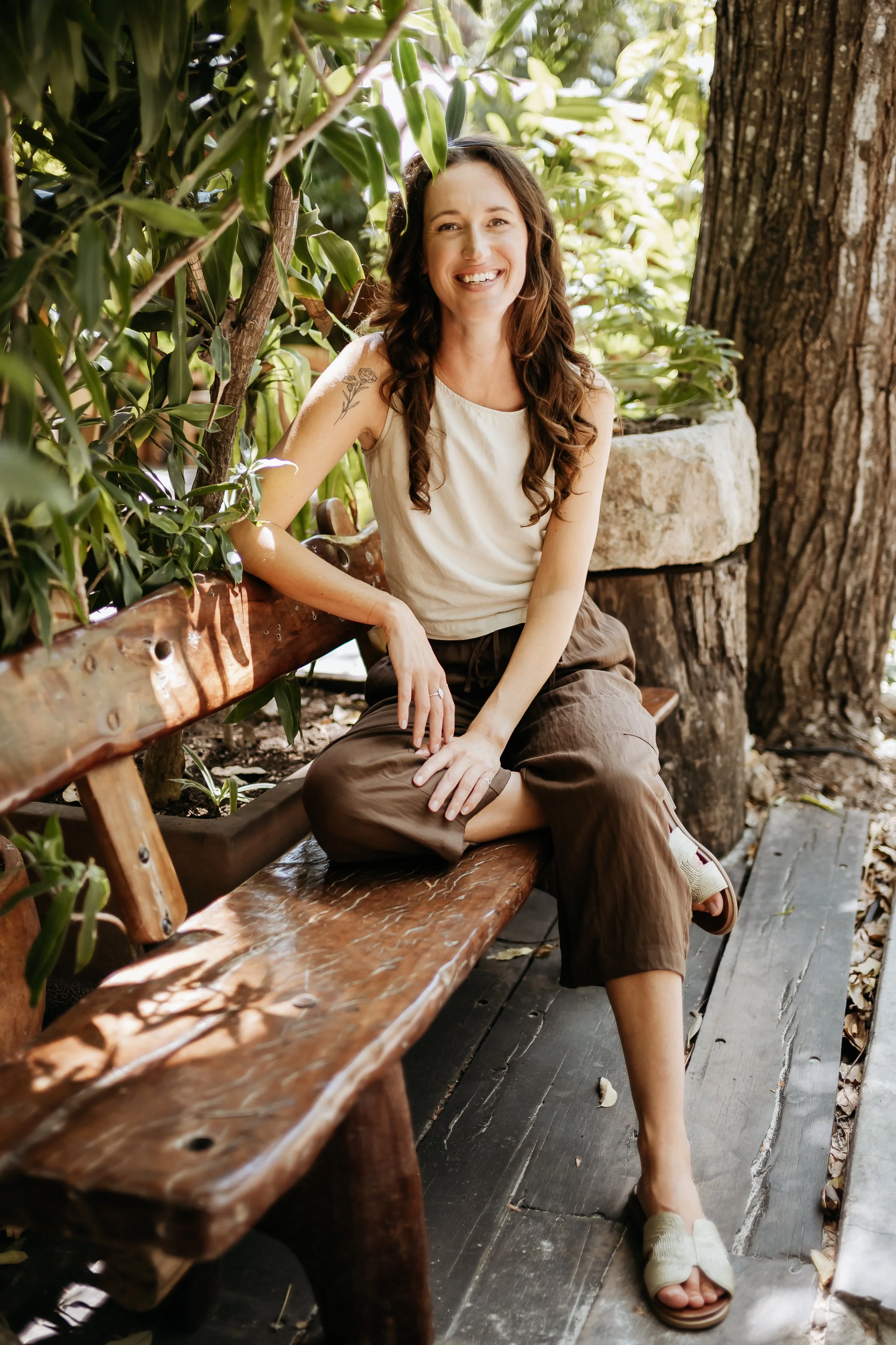 Woman sitting on white steps, smiling, wearing a gray tank top and brown shorts, with leafy plants nearby.