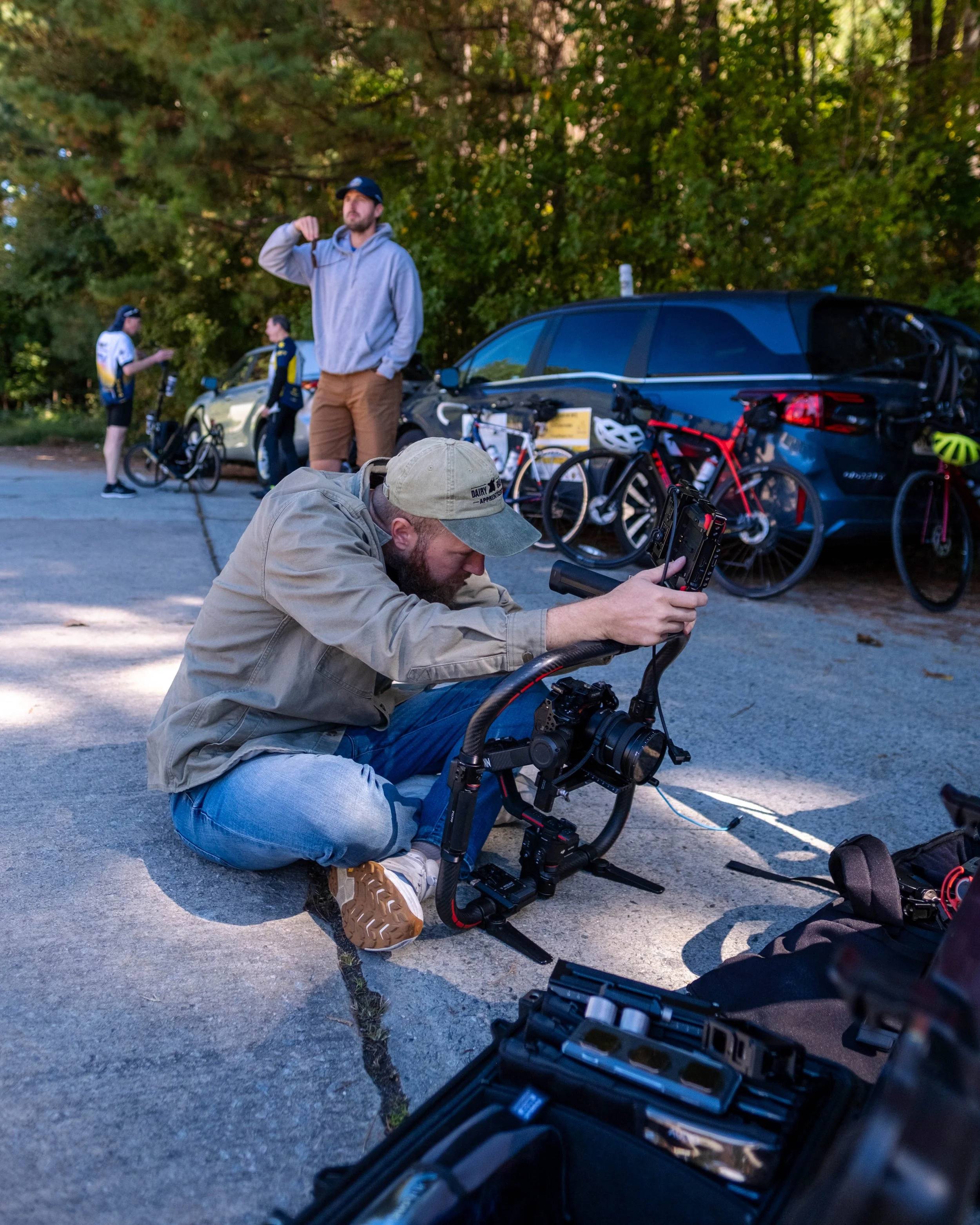 Two men sitting on a red utility vehicle outdoors in a green field with trees in the background. One man is holding a remote control, and the other is operating a camera mounted on a stabilizer rig, both wearing hats and smiling.