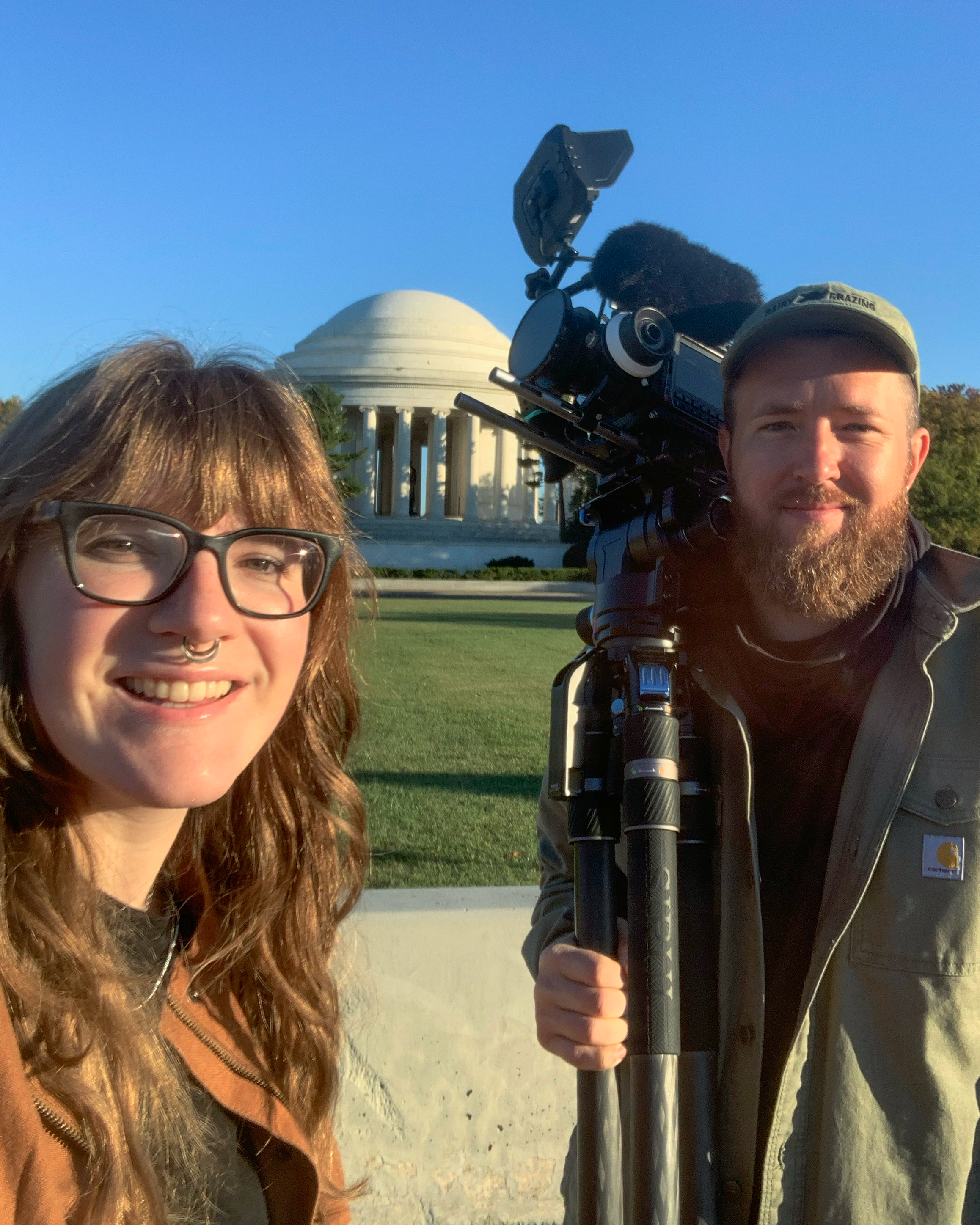 Two people stand in front of the Lincoln Memorial at sunset, one person is a woman with glasses and a septum piercing, the other is a man with a beard and cap, holding a professional video camera on a tripod.