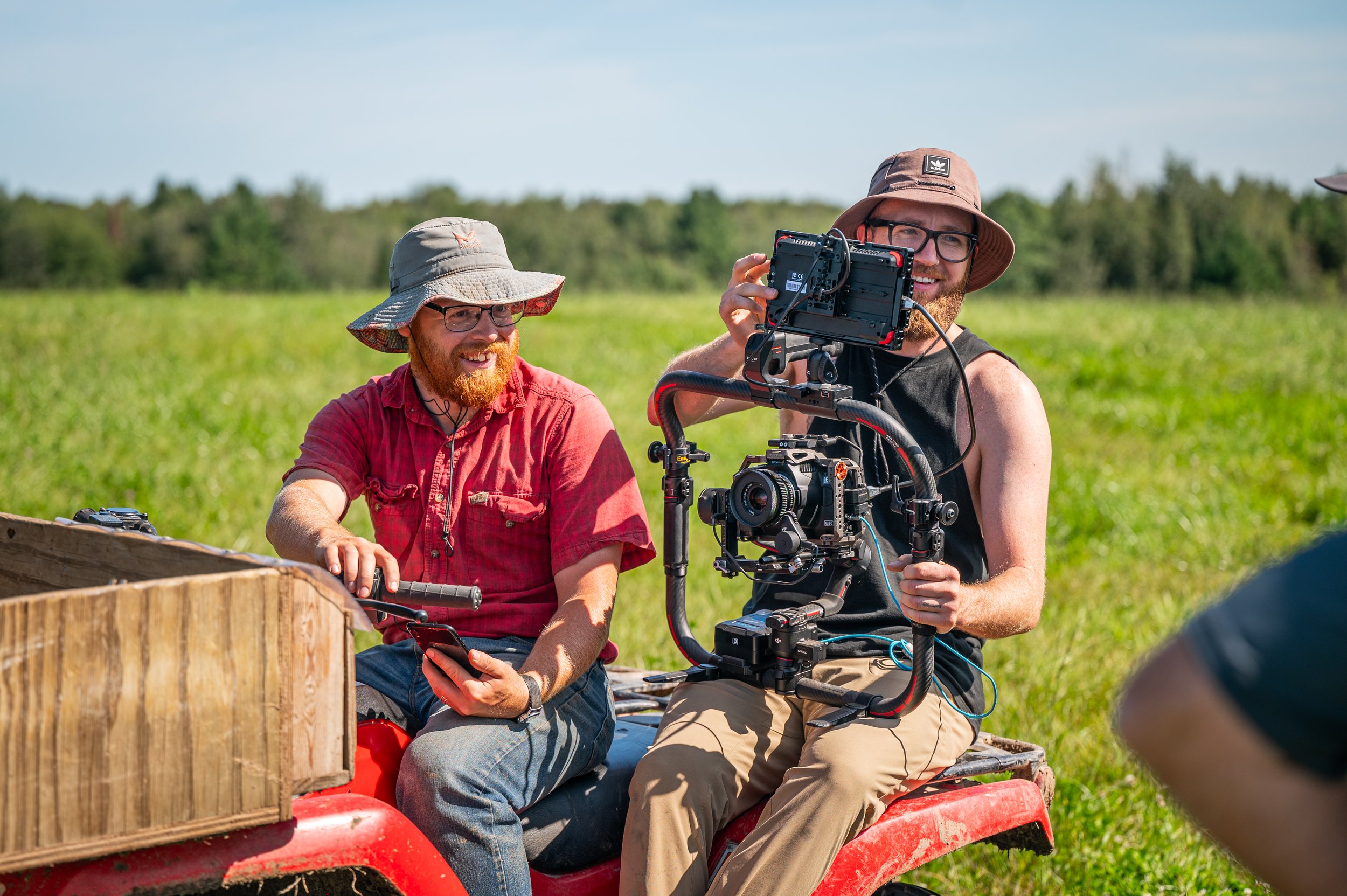 Two men sitting on a red utility vehicle outdoors in a green field with trees in the background. One man is holding a remote control, and the other is operating a camera mounted on a stabilizer rig, both wearing hats and smiling.