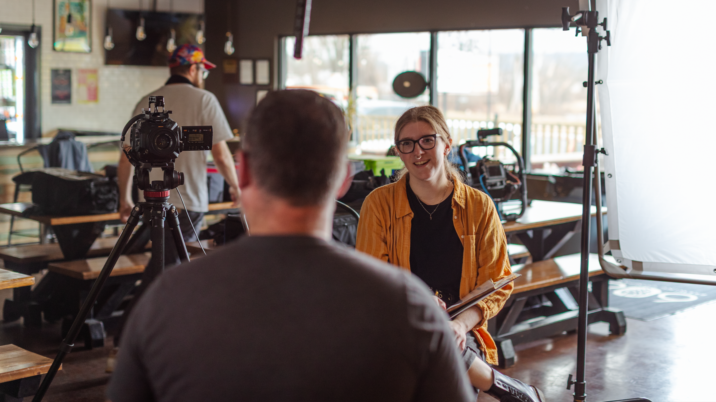 A young woman with glasses, wearing a yellow jacket, is being interviewed in a cafe or restaurant. She is smiling and holding a notepad. There is a camera on a tripod filming her, and a man with short hair, seen from behind, is conducting the interview. Large windows with natural light are in the background.