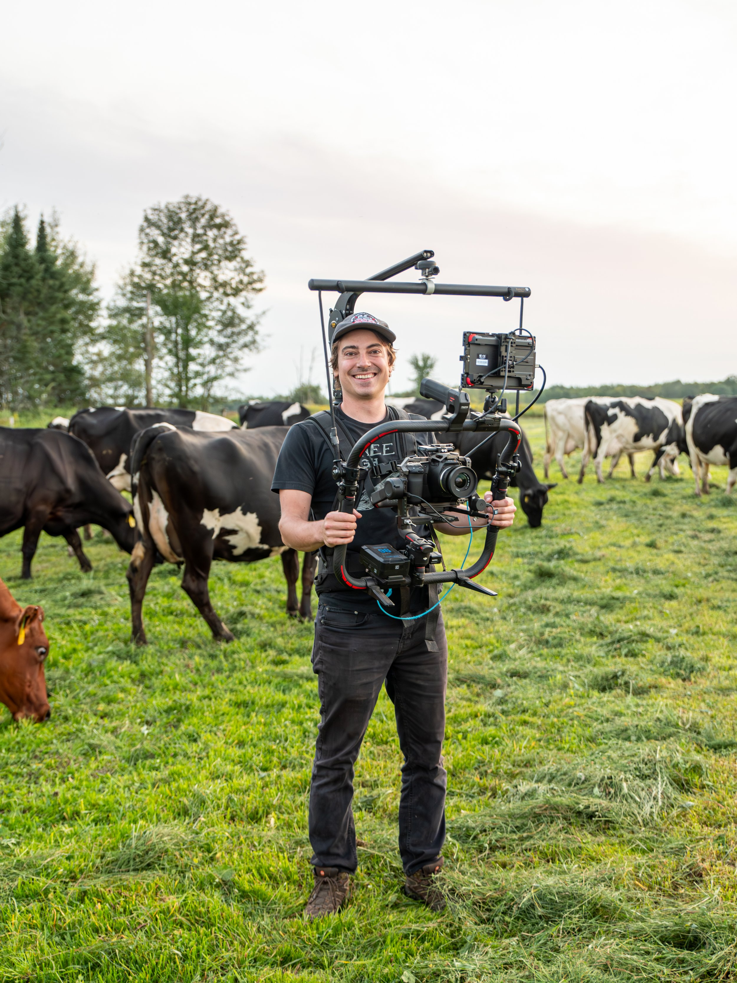 A smiling man in black clothes and a cap holding a camera rig in a field with cows grazing.
