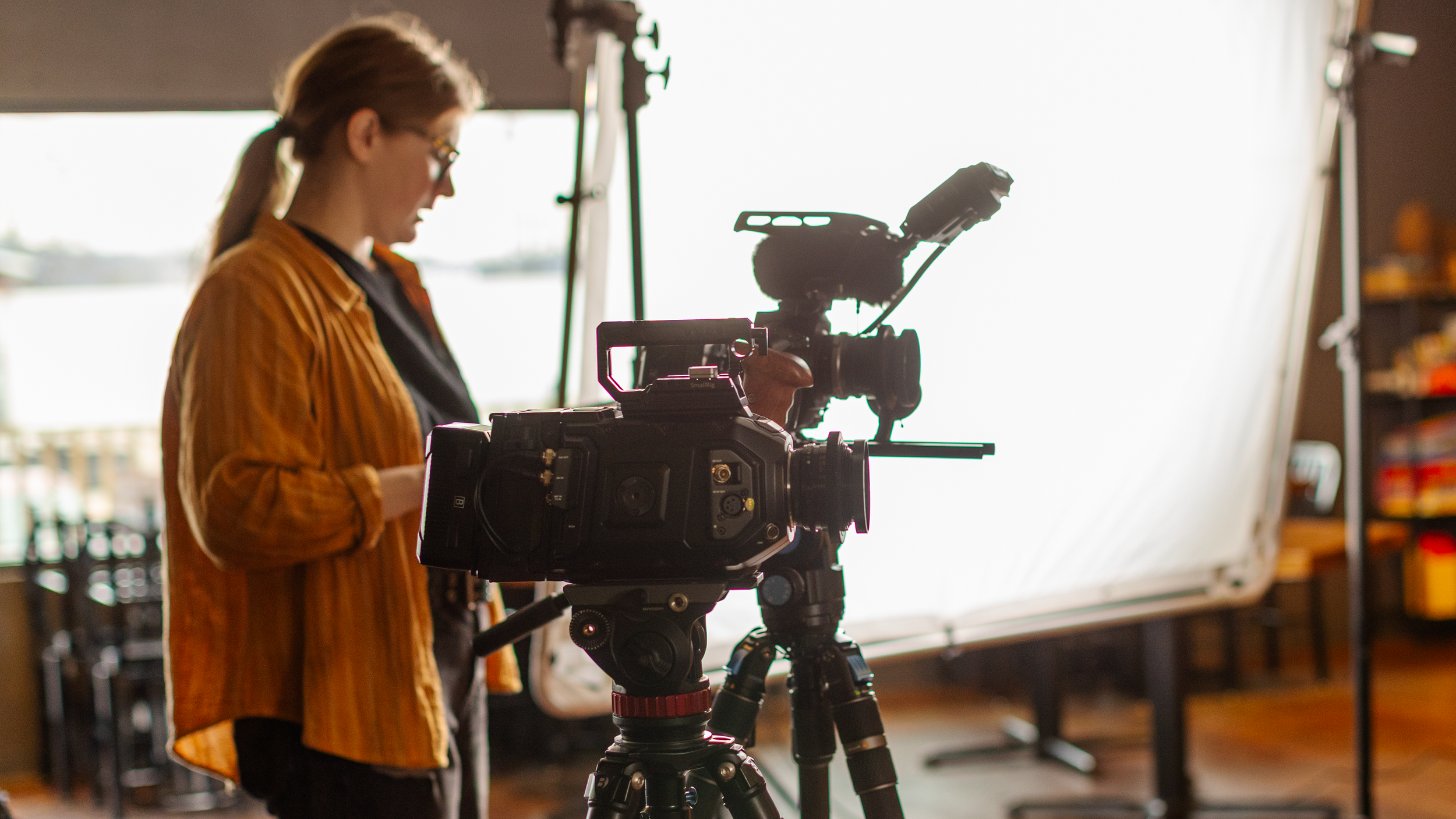 A woman operating a professional video camera in a studio setting with lighting equipment and a large white backdrop.