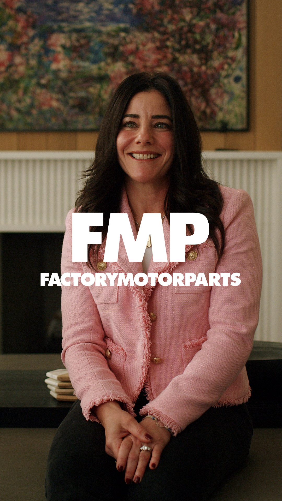 A woman with dark hair smiling, wearing a pink jacket, sitting in front of a colorful painting.