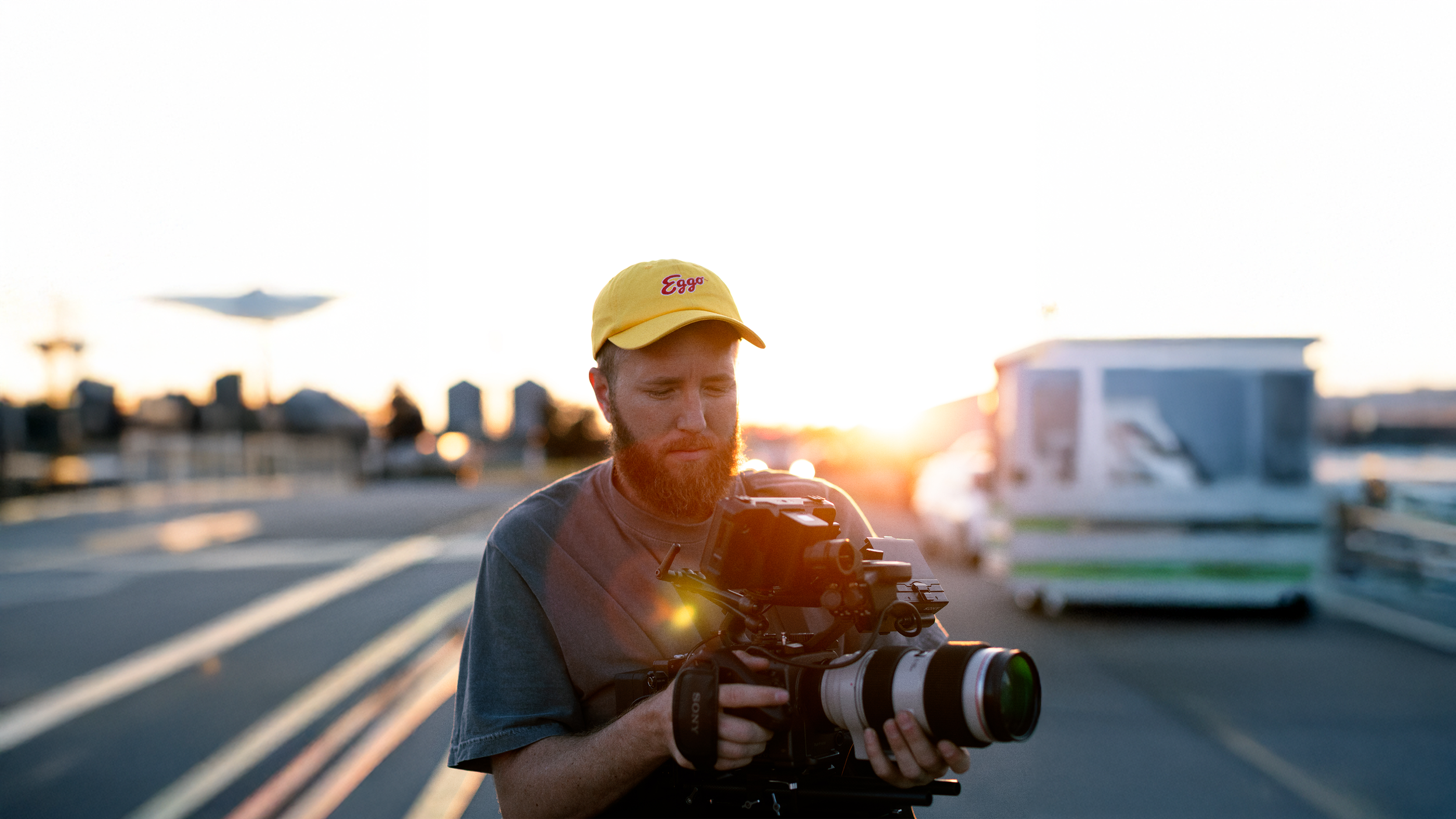 Man with a red beard wearing a yellow cap with 'Eggs' written on it, holding a professional camera with a telephoto lens, standing outdoors on a bridge at sunset.