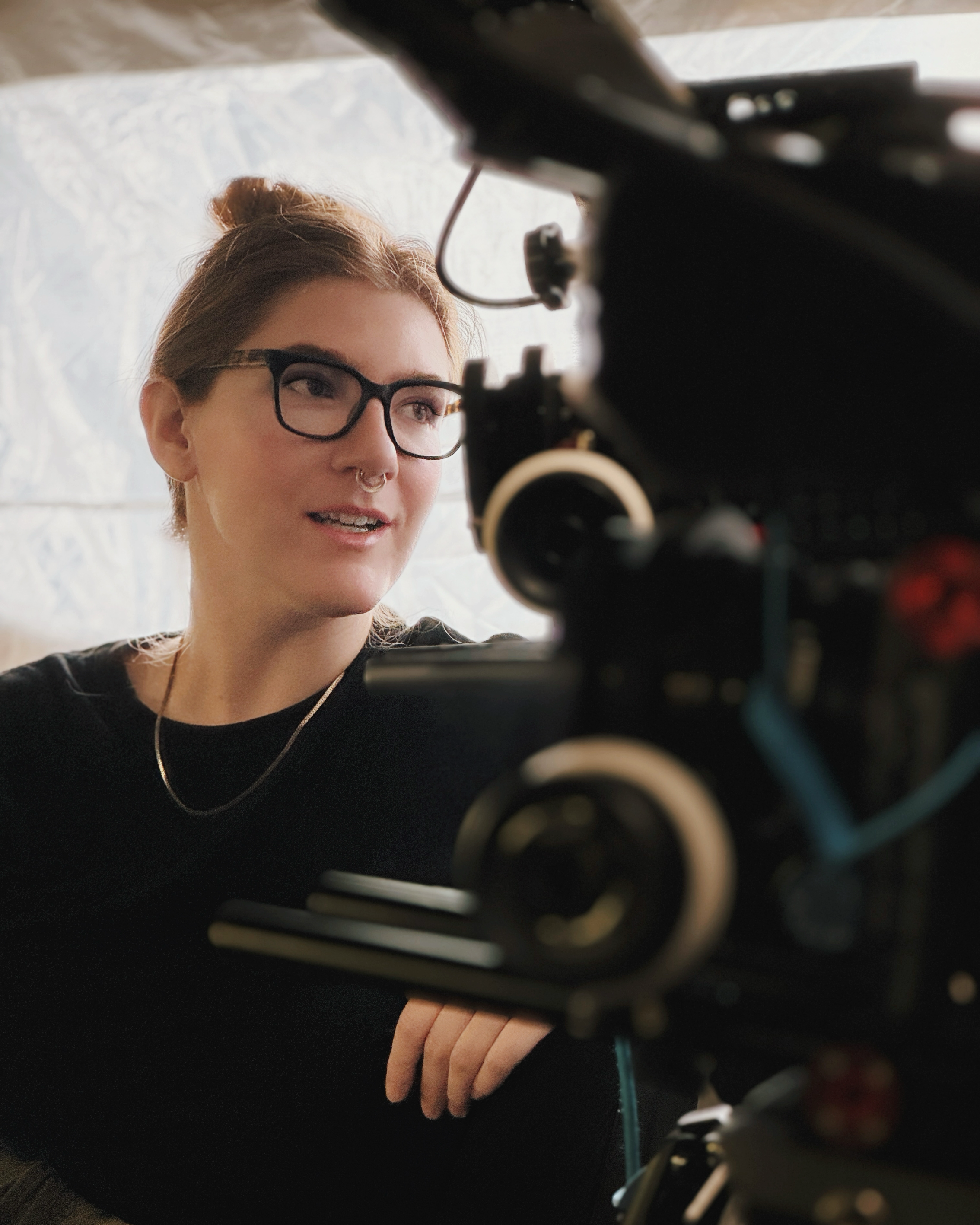A woman with glasses and a septum piercing looks at a film camera during a shoot.