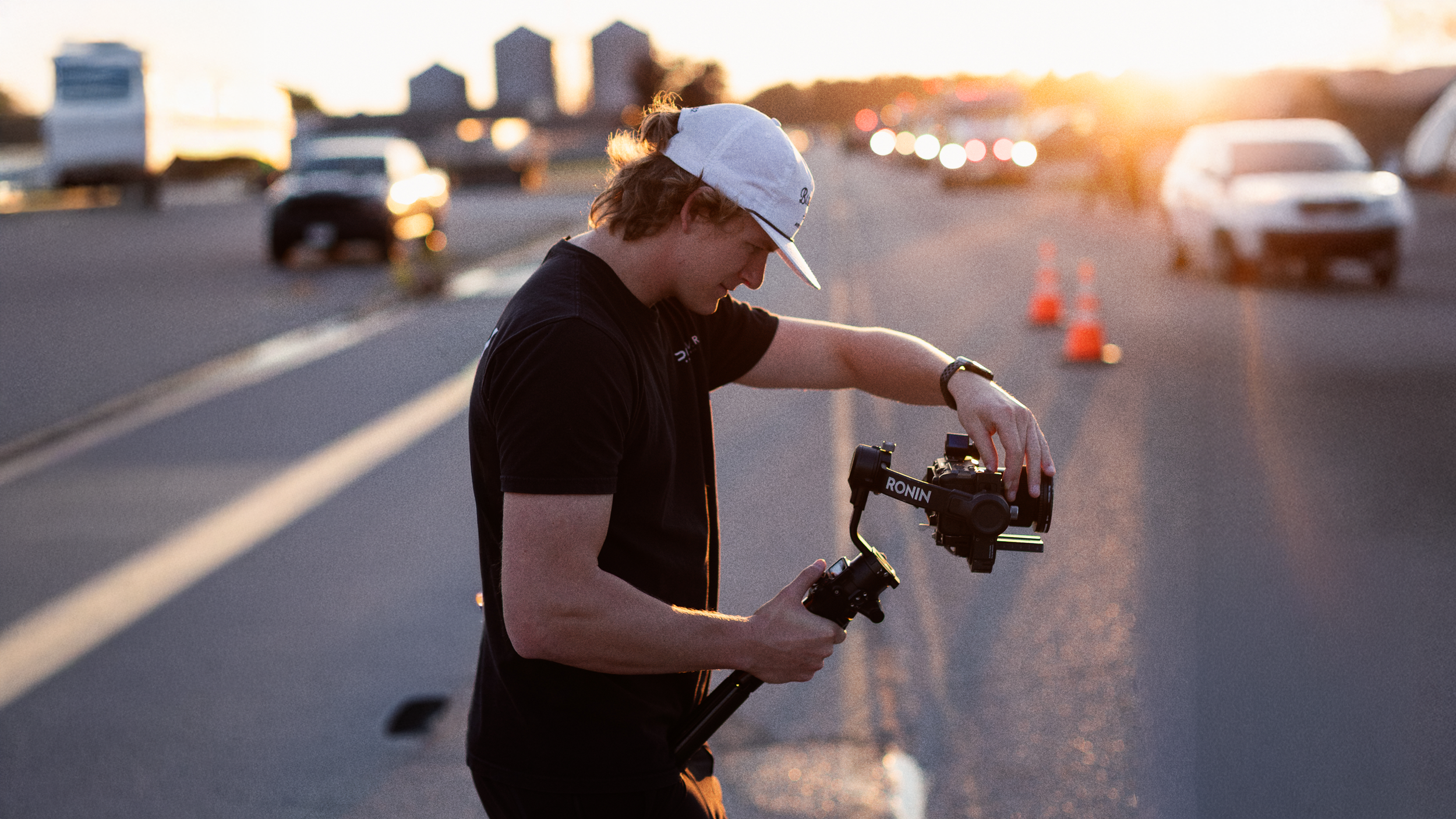 A young man with a white cap and black T-shirt setting up a camera with a stabilizer on a road during sunset, with cars and traffic cones in the background.