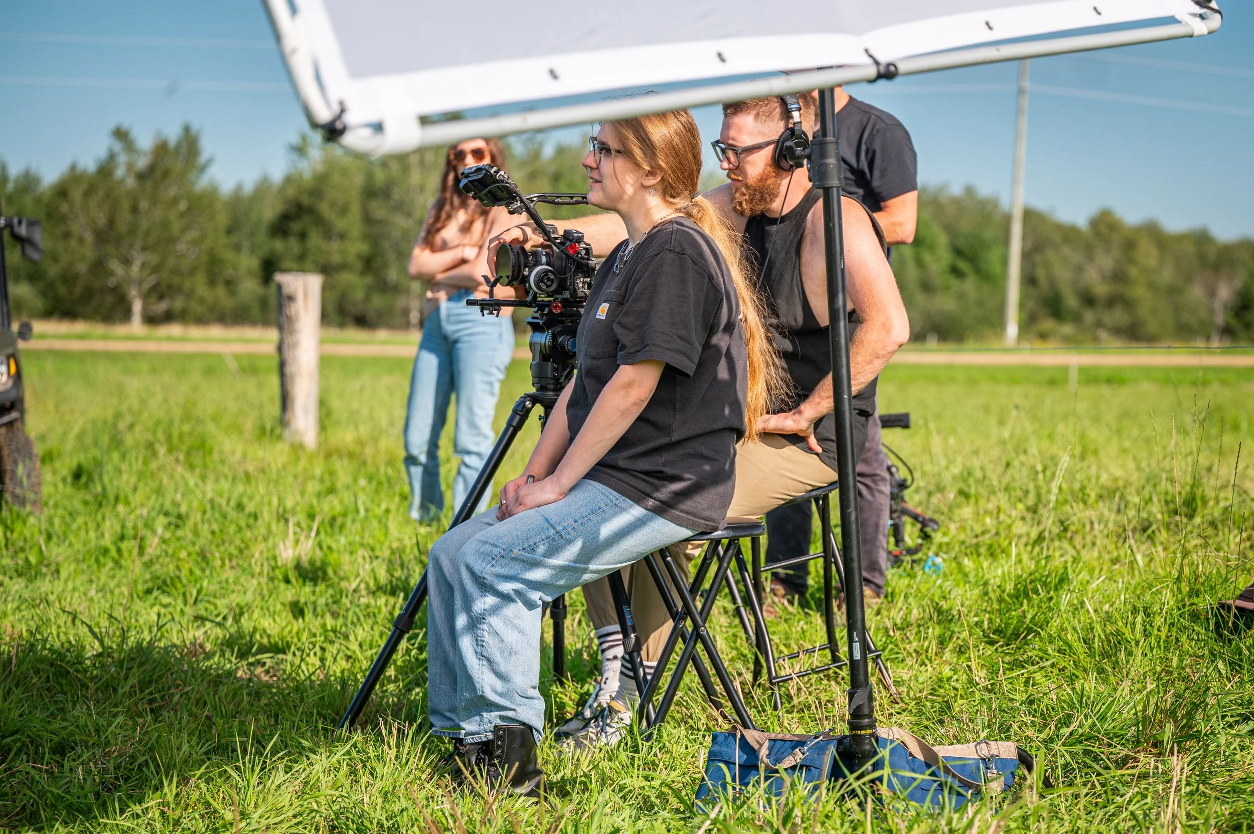 Film crew on outdoor set with camera operator and director during daytime, surrounded by green grass and trees.