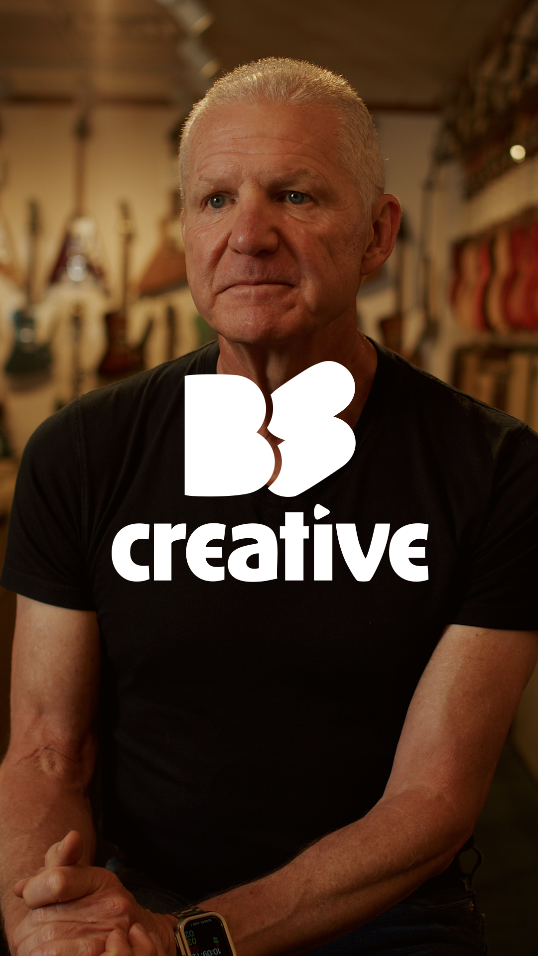 A mature man with short gray hair and a black t-shirt, sitting with arms crossed, in a room with guitars hanging on the wall, and the logo 'BE creative' overlayed on the image.