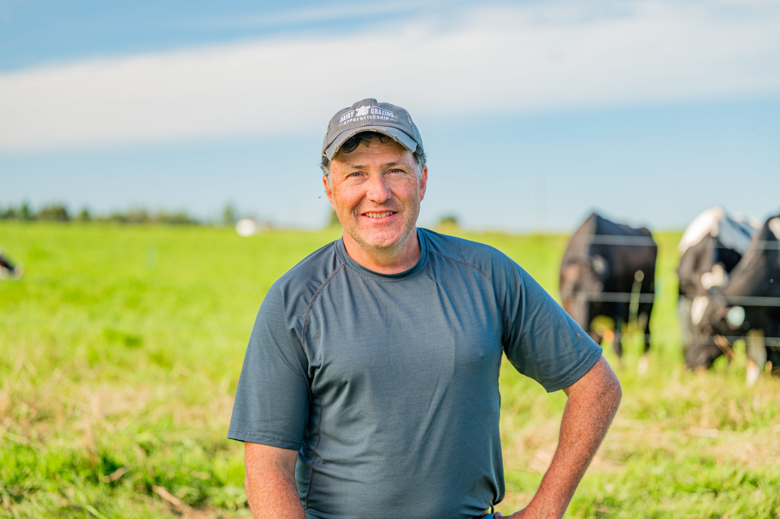 Smiling middle-aged man in a gray athletic shirt and cap standing in a lush green field with cows grazing in the background under a partly cloudy sky.