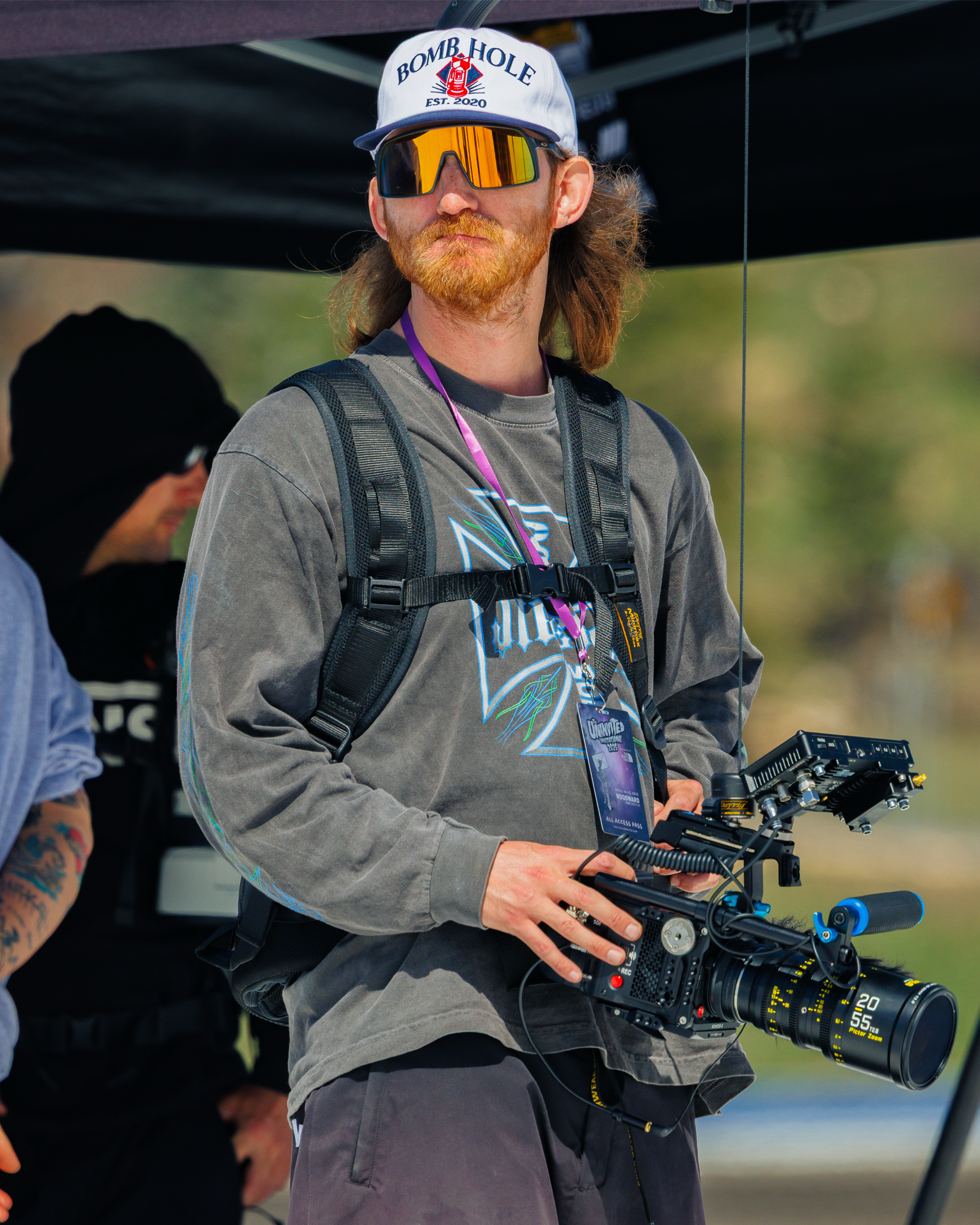A man in sunglasses, a baseball cap that reads 'BOMB HOLE' with an illustration of a bomb, and a gray sweatshirt, is holding a professional camera with a large lens. He is wearing a backpack and an event pass around his neck, standing outdoors.