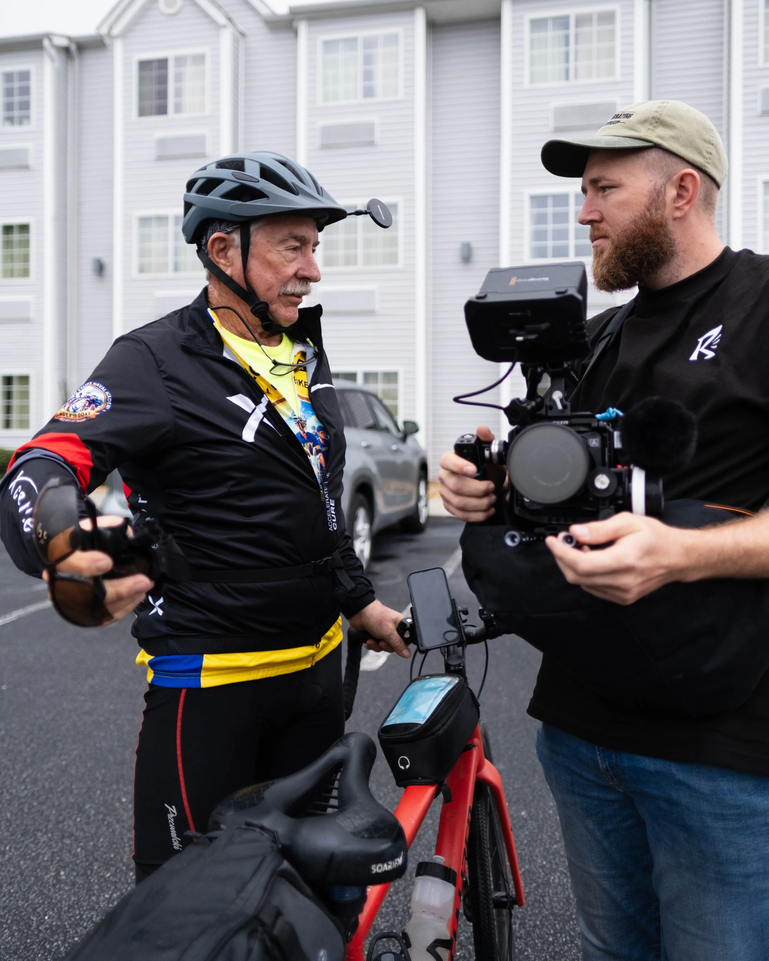 A smiling man in black clothes and a cap holding a camera rig in a field with cows grazing.