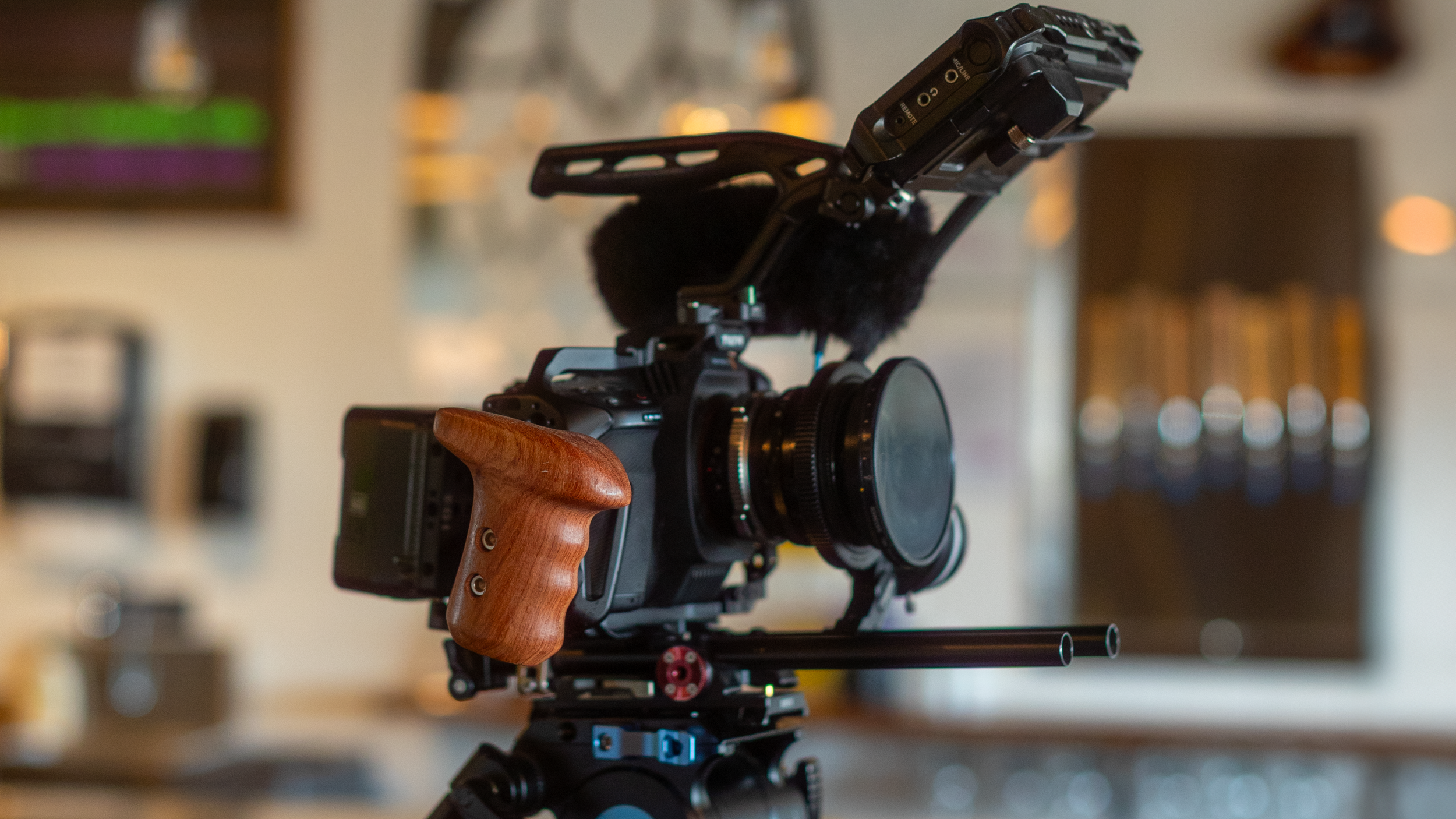 Close-up of a professional video camera with a wooden handle, mounted on a tripod, with a microphone attached on top. The background is blurred, indicating an indoor setting.