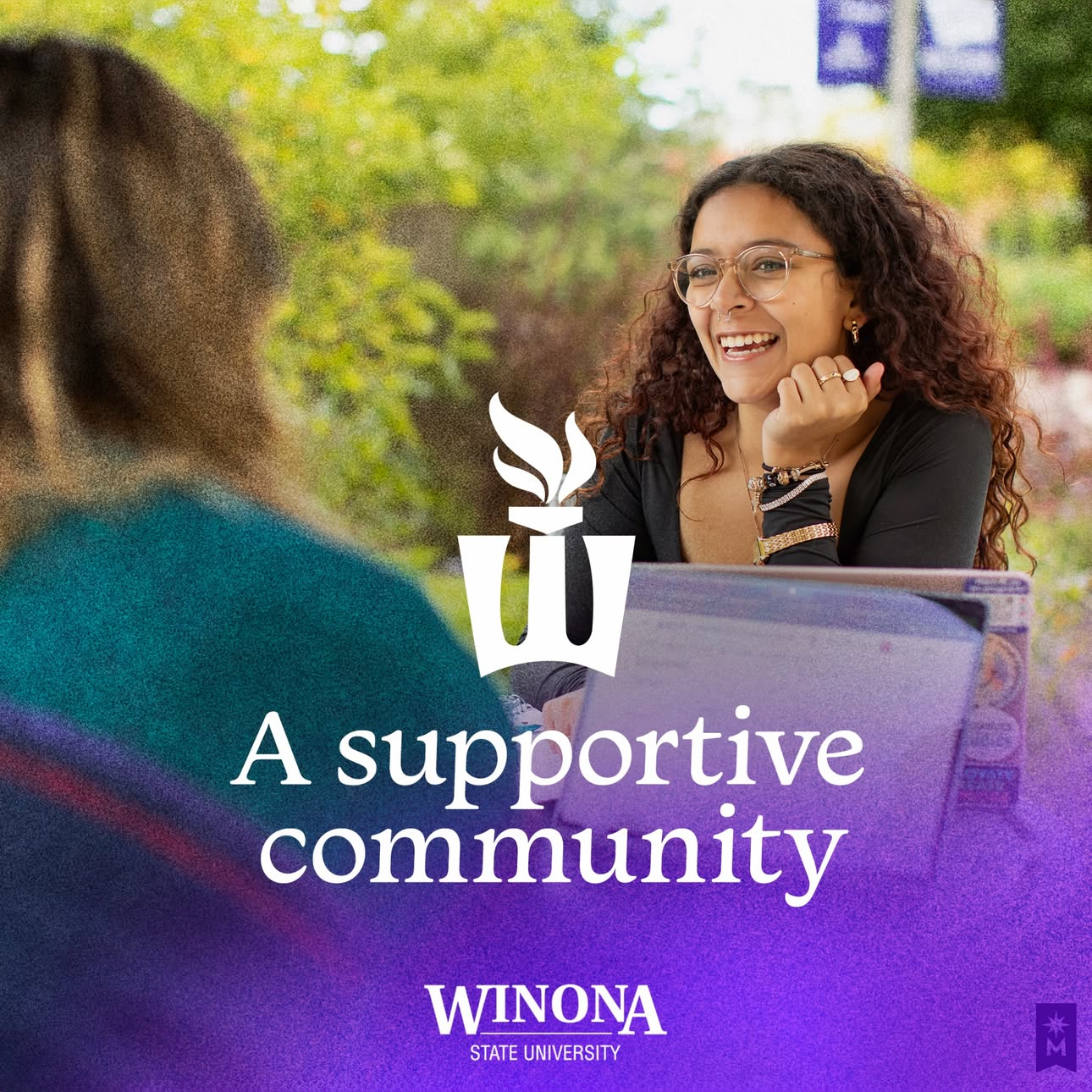 A smiling woman with curly hair and glasses talking to another person at an outdoor table, with trees in the background, promoting a supportive community at Winona State University.