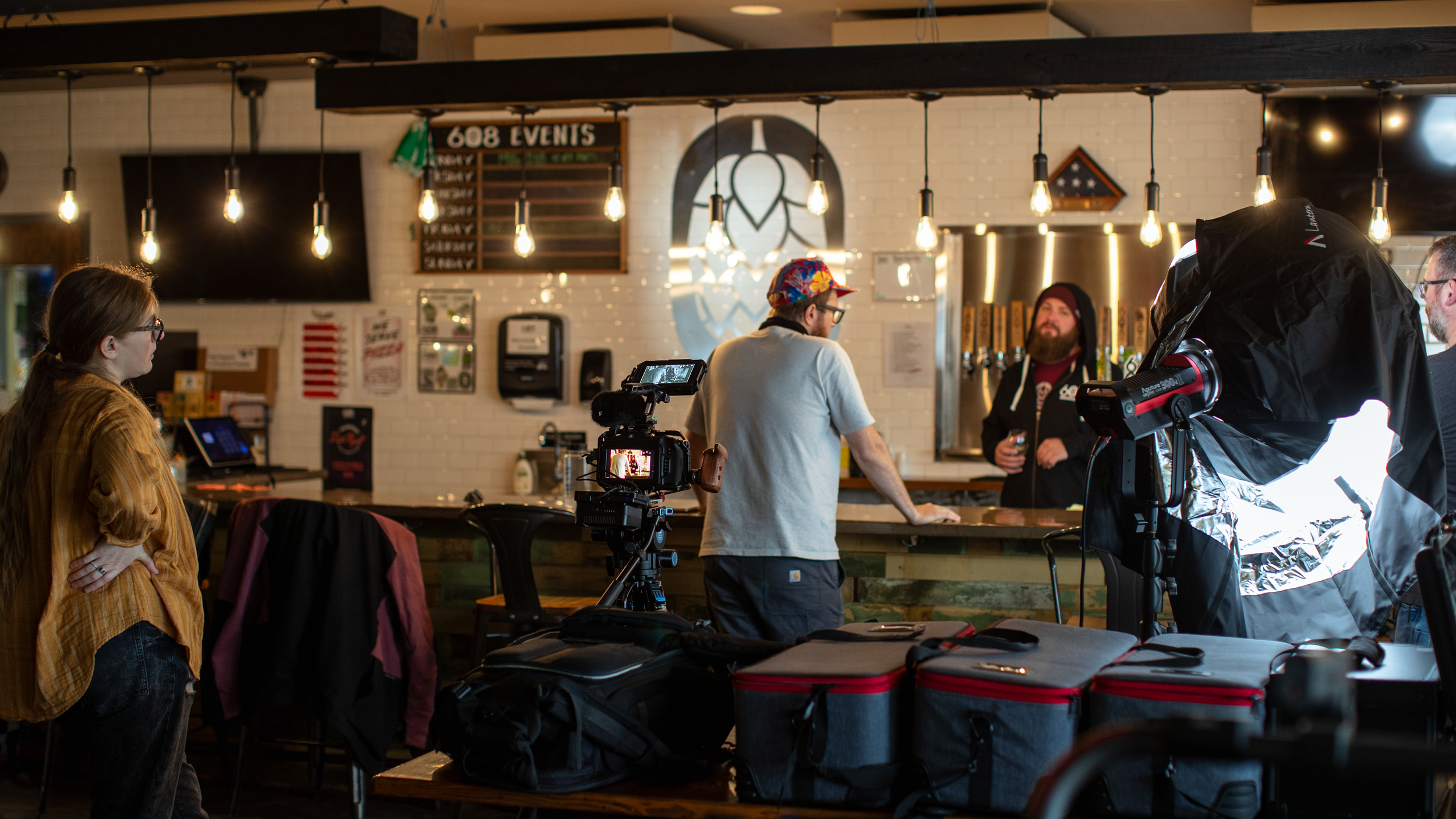 Filming crew setting up camera and lighting in a bar or brewery, with a woman observing, multiple people behind the counter, and ambient hanging lights.