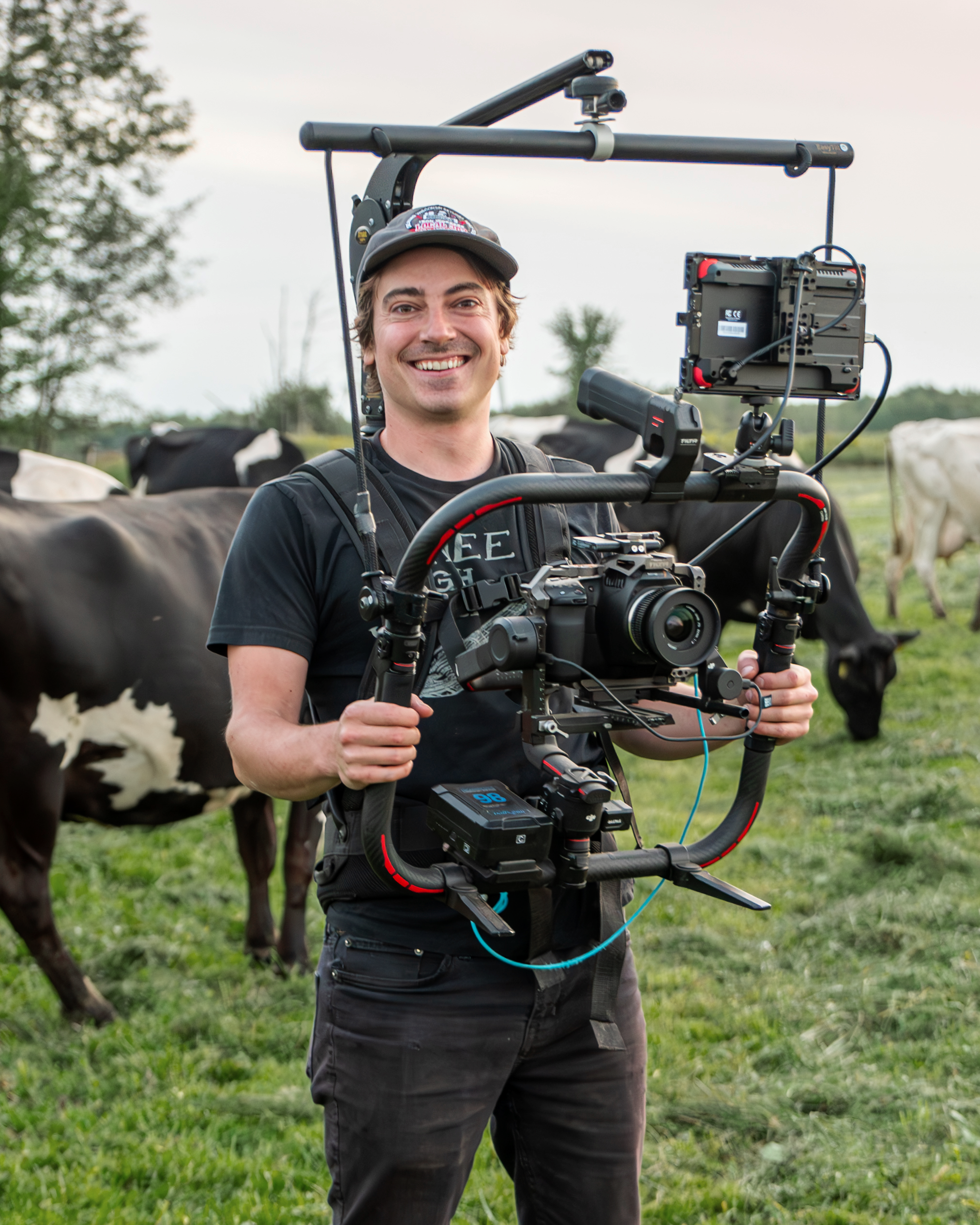 A man operating a professional camera rig outdoors in a field, with cows grazing in the background, smiling at the camera.