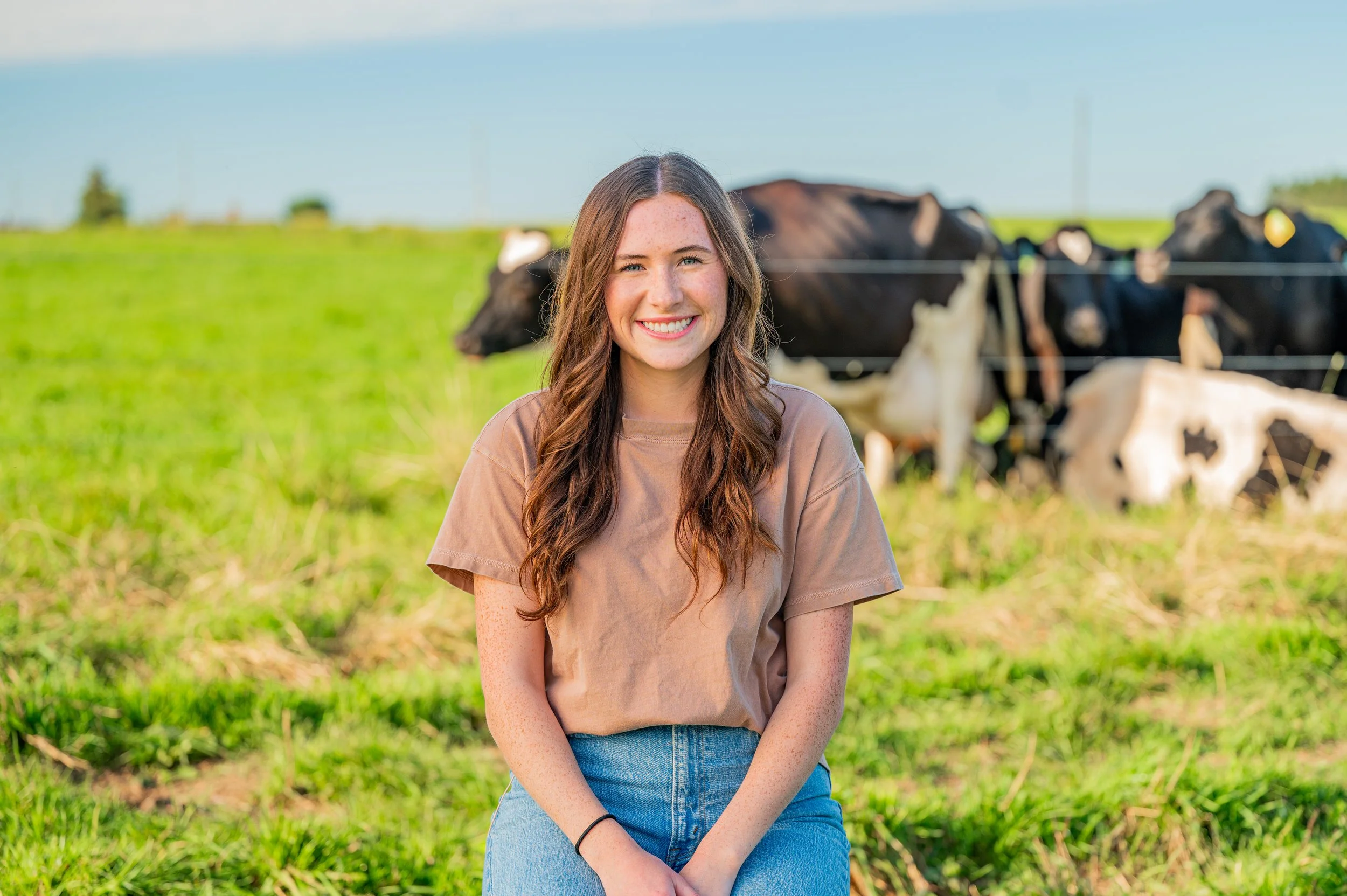 A young woman with long brown hair, smiling, wearing a beige t-shirt and blue jeans, sitting on grass in a field with cows in the background.