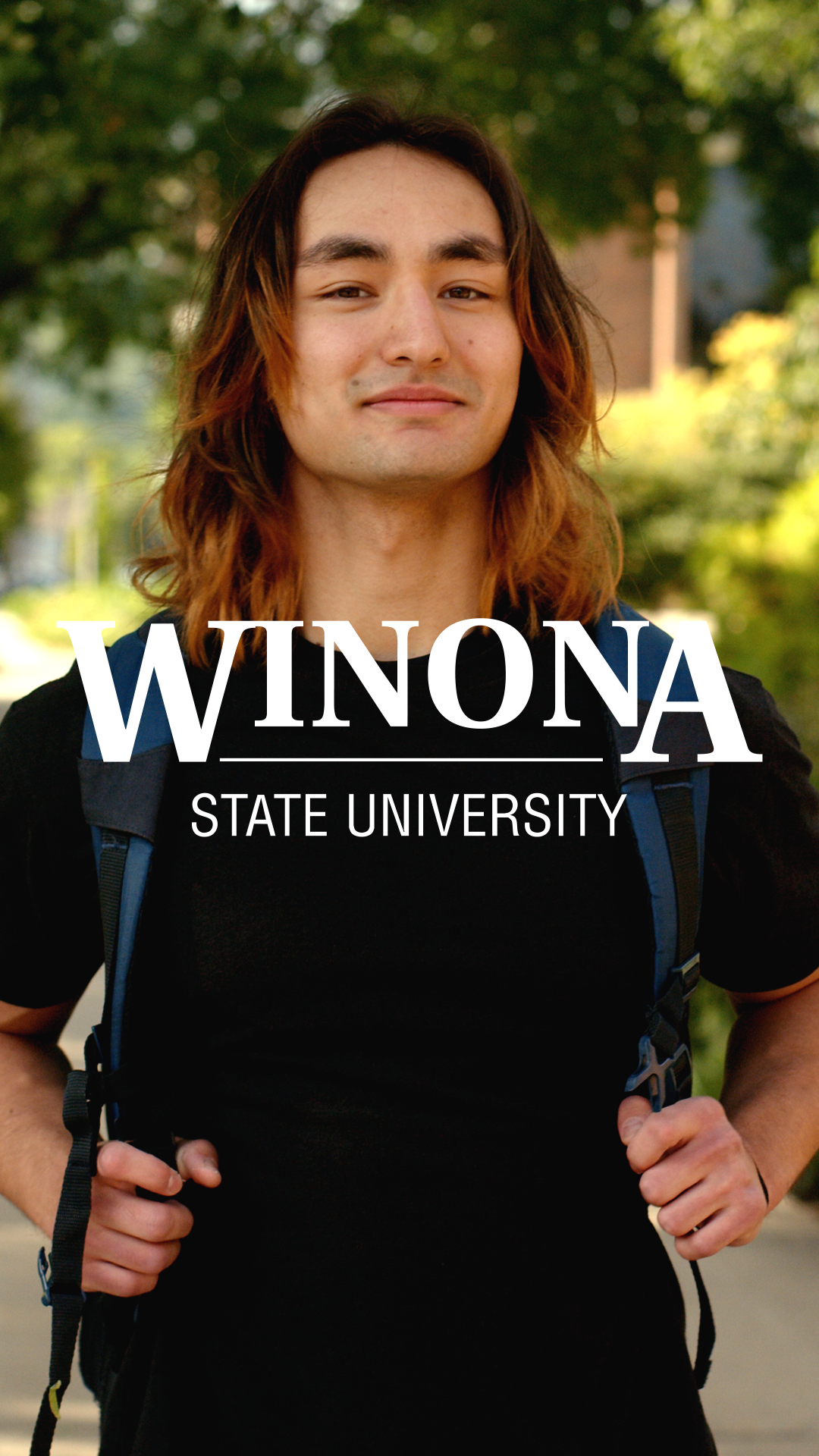 A young man with long brown hair standing outdoors with green trees in the background. He is wearing a black shirt and carrying a blue backpack. The text 'Winona State University' is overlaid on the image.