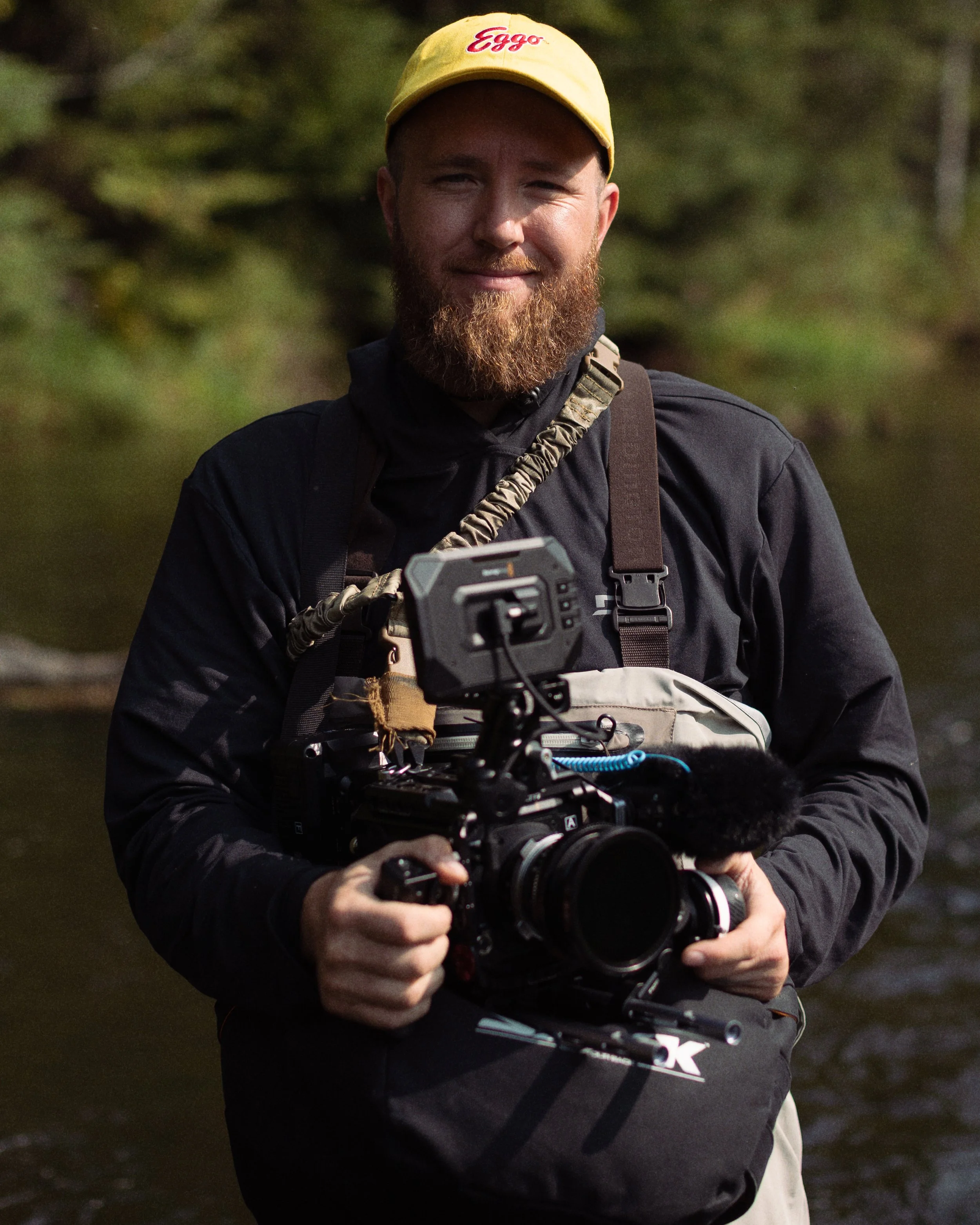 A man with a beard wearing a yellow hat, black jacket, and carrying a camera setup outdoors near a river, smiling at the camera.