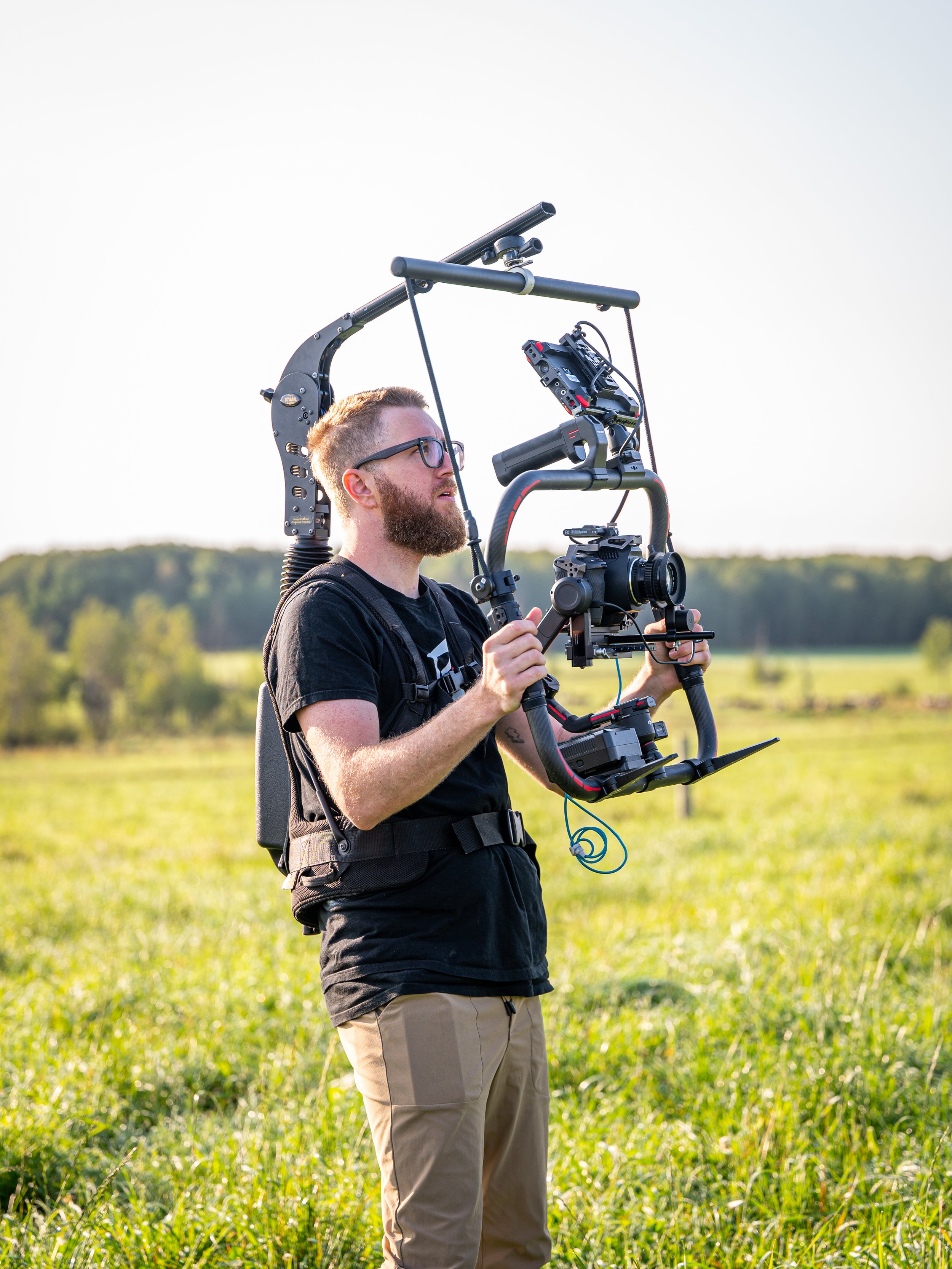 A man with glasses and a beard is filming outdoors using a professional camera stabilizer rig, standing on a grassy field with trees in the background.