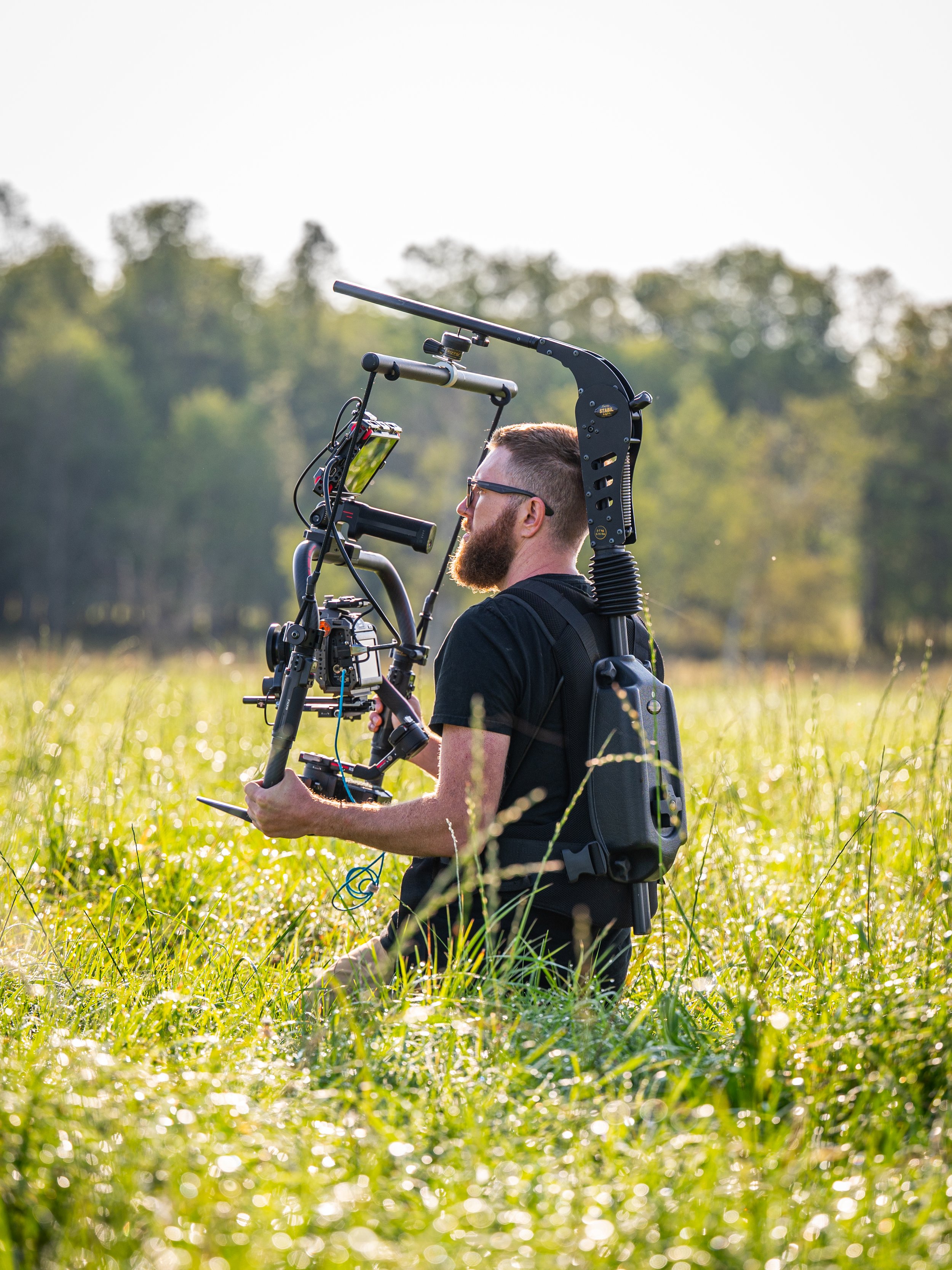 A man with a beard and glasses kneels in a grassy field holding a camera stabilizer rig.