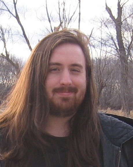 A man with long brown hair and a beard, smiling outdoors during winter with leafless trees in the background.