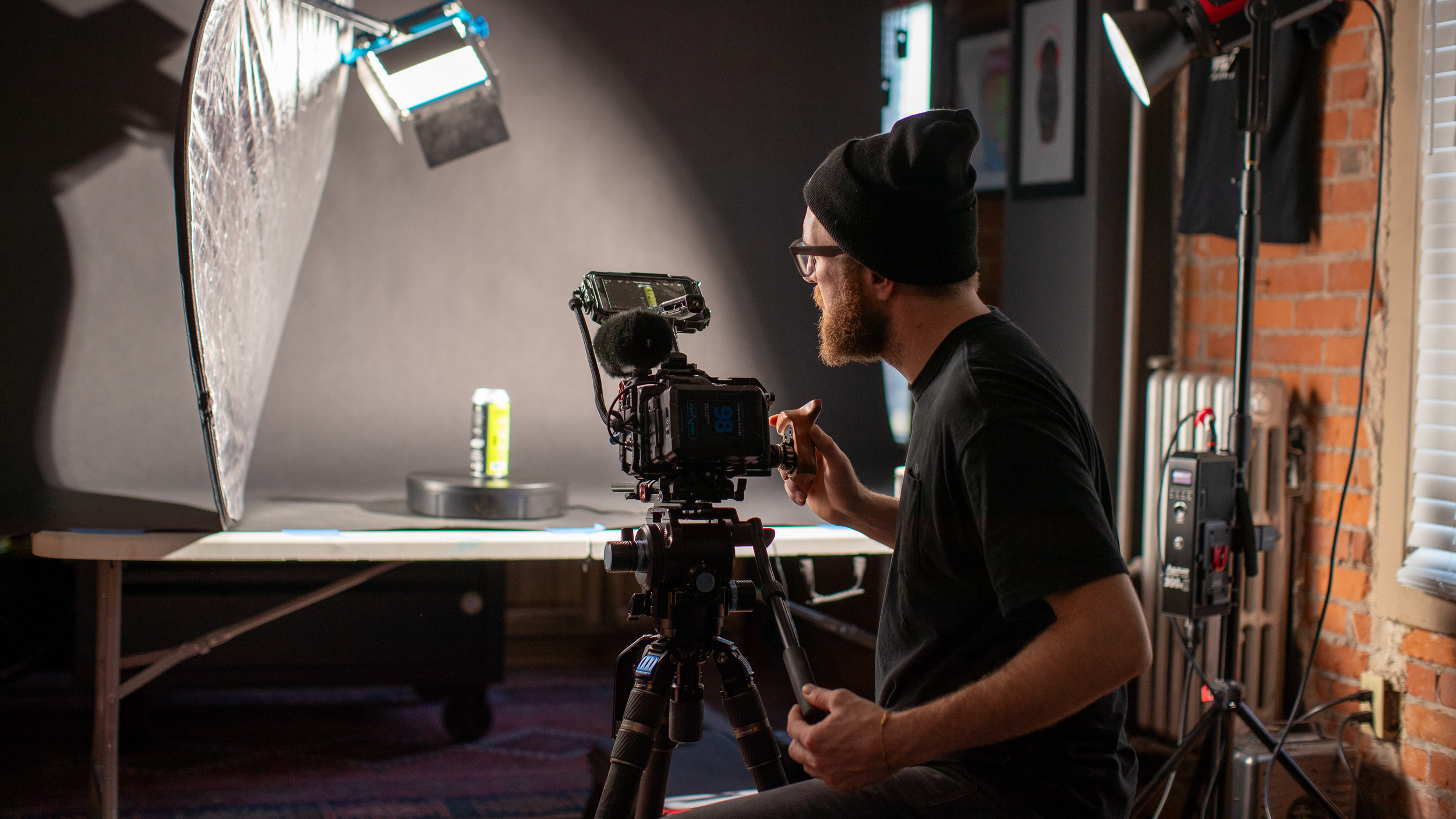 A man with a black beanie, glasses, and a beard operating a professional video camera in a photography studio set with props, lighting, and a gray backdrop, creating a product shot of a can.