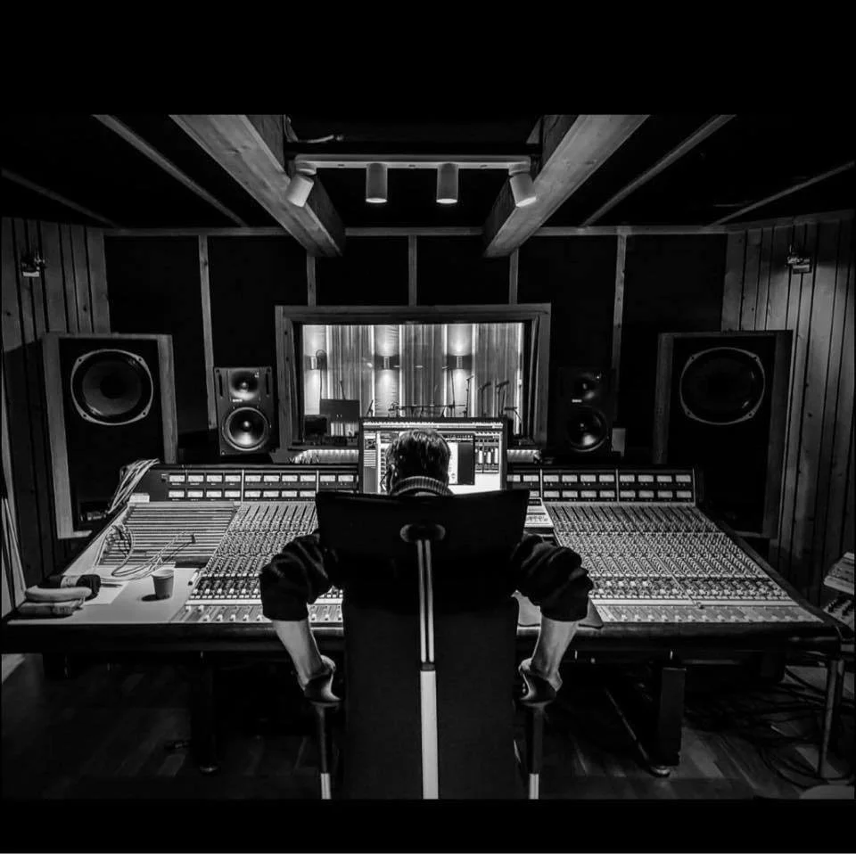 A music recording studio with a person working at a large mixing console, surrounded by speakers, with a window showing a recording booth in the background.