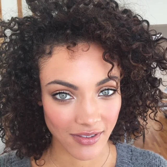 A young woman with curly dark hair, bright blue eyes, and a subtle smile. She is wearing a gray top and a delicate necklace, with a blurred indoor background.