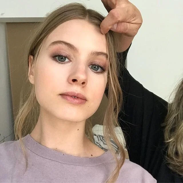 A young woman with long blonde hair and blue eyes looks into the camera, with a person's hand touching her forehead. The woman is wearing a light purple shirt and has makeup on, including foundation, eyeshadow, and lipstick.
