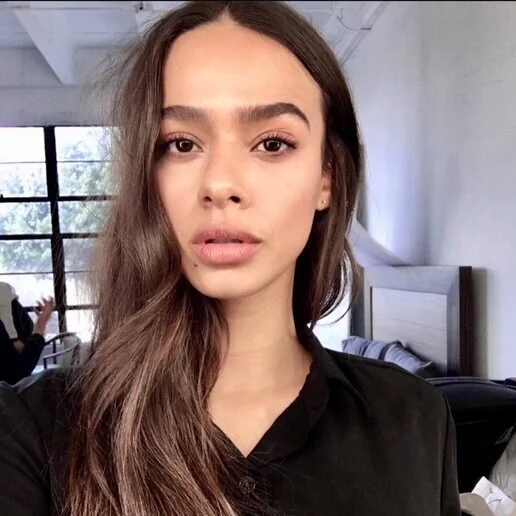Young woman with long brown hair and full eyebrows taking a selfie in a bright room with large windows, a bed, and a person sitting at a desk in the background.
