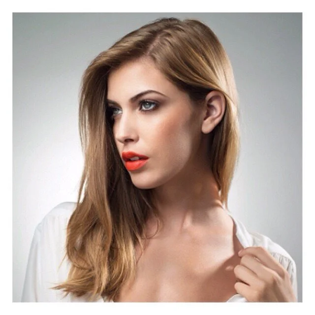 A young woman with long, light brown hair and blue eyes, wearing red lipstick and a white top, looking to the side against a neutral background.