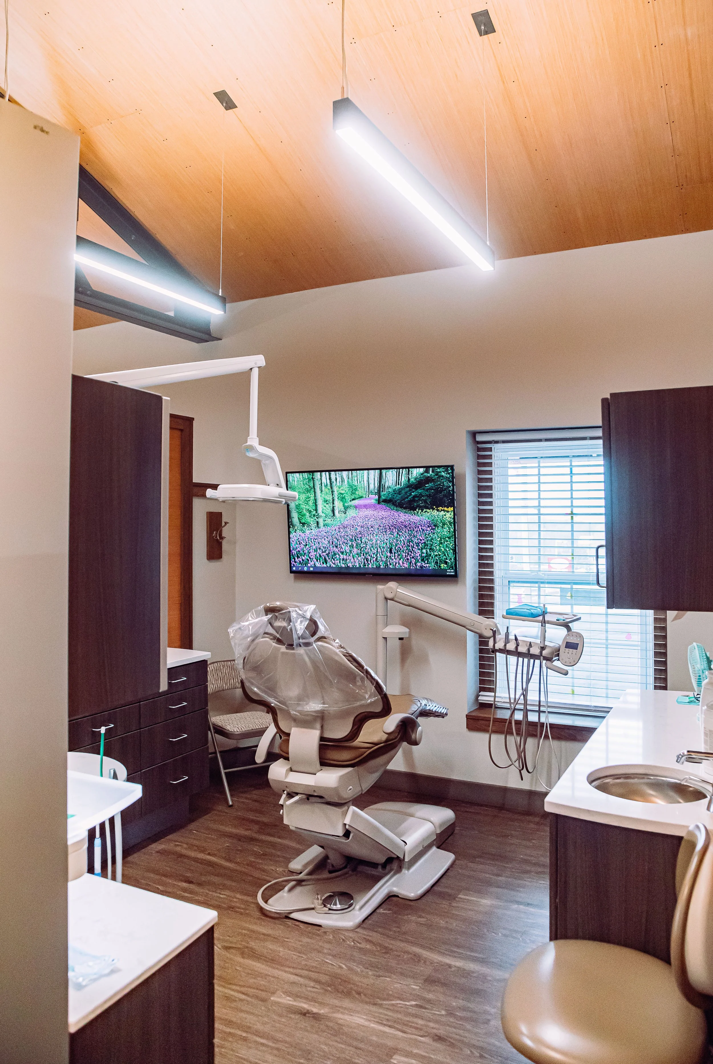 Very clean and organized dental examination room in cream and brown with cabinets, a TV, and top-of-the-line dental equipment