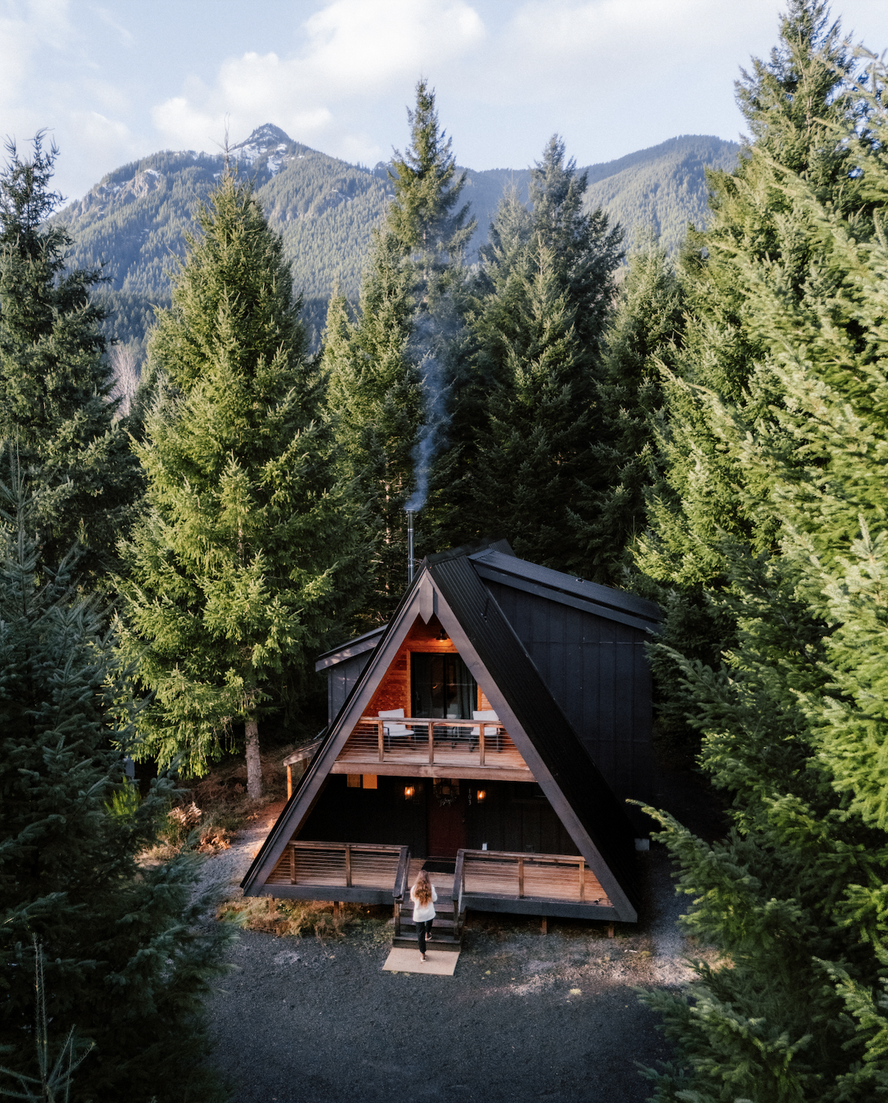 A modern A-frame house with a balcony, surrounded by tall green trees.
