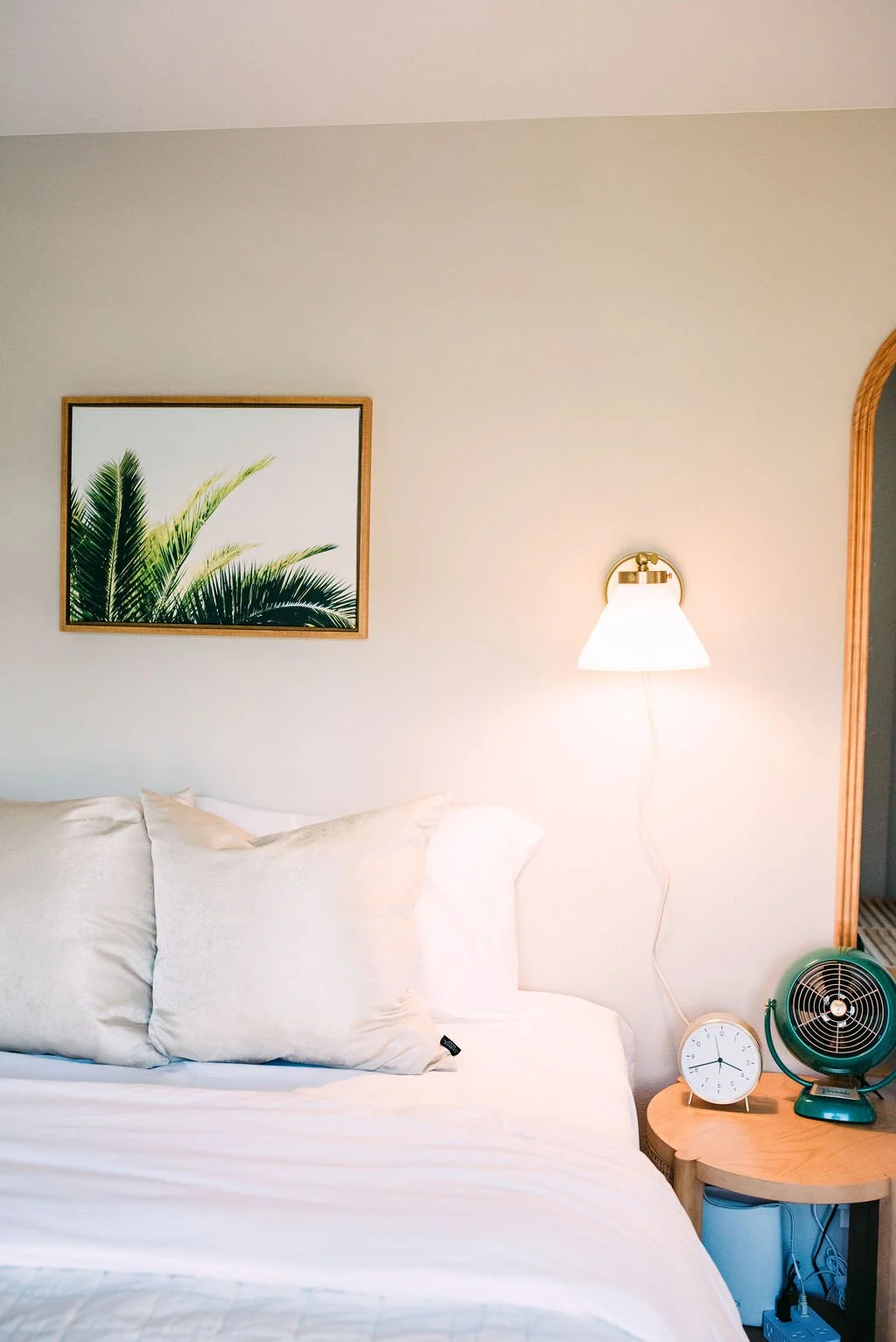 A bedroom corner with a white bed, beige pillows, a wall-mounted lamp, a framed palm leaf print, a small wooden nightstand with a clock and a small fan, and an arched wooden frame on the right.